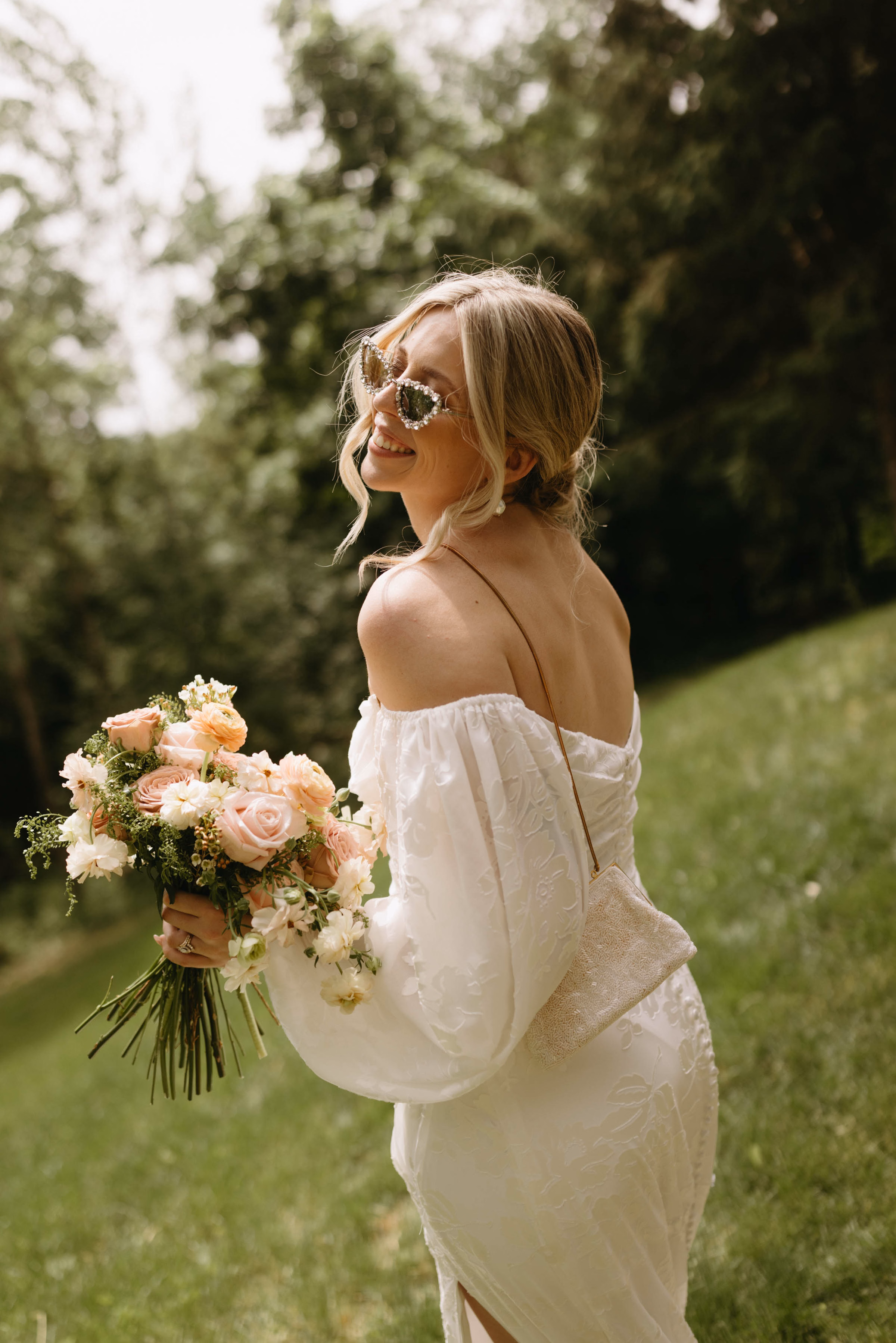 A smiling bride in a modern lace gown with puff sleeves wearing pearl-embellished sunglasses, with her day perfectly styled by Skies the Limit Events, a wedding planner and party planner in La Crosse, WI. She carries a romantic, custom-designed bridal bouquet featuring soft peach and pink roses mixed with delicate white sweet peas.
