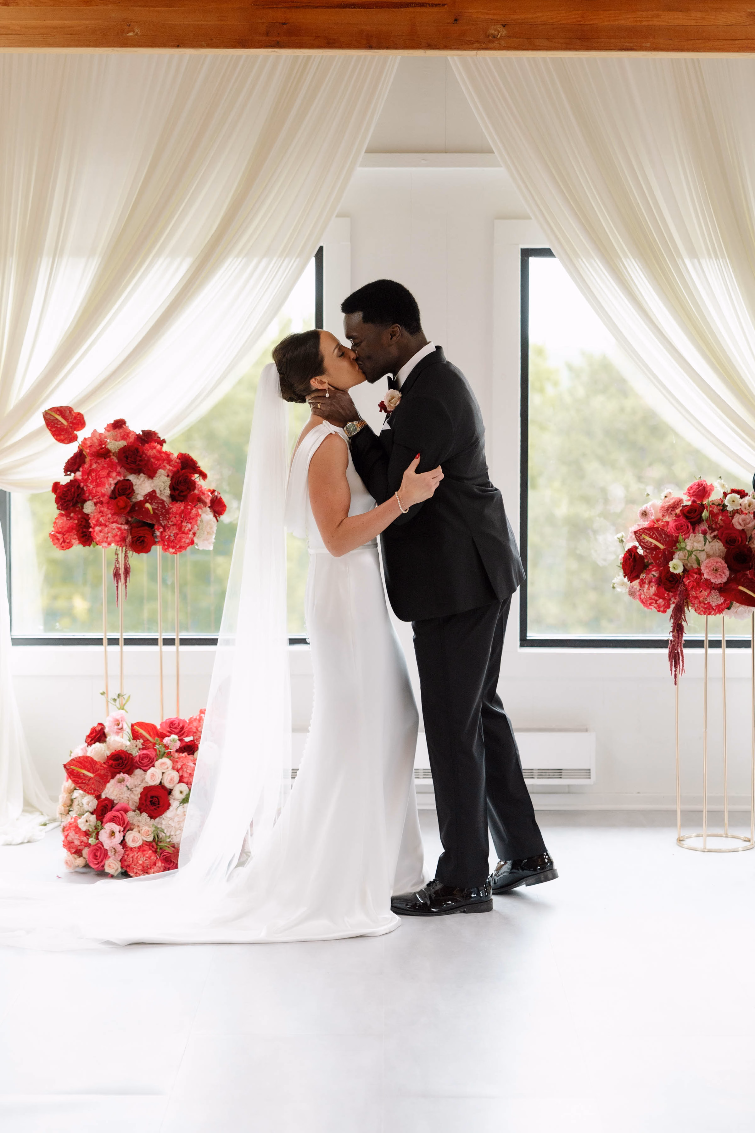 A bride and groom sharing their first kiss under soft white drapery, planned and designed by Skies the Limit Events, a wedding planner in La Crosse, WI. The modern indoor ceremony space is framed by her custom floral installations featuring striking shades of red and pink roses, carnations, and anthuriums on elegant gold stands.