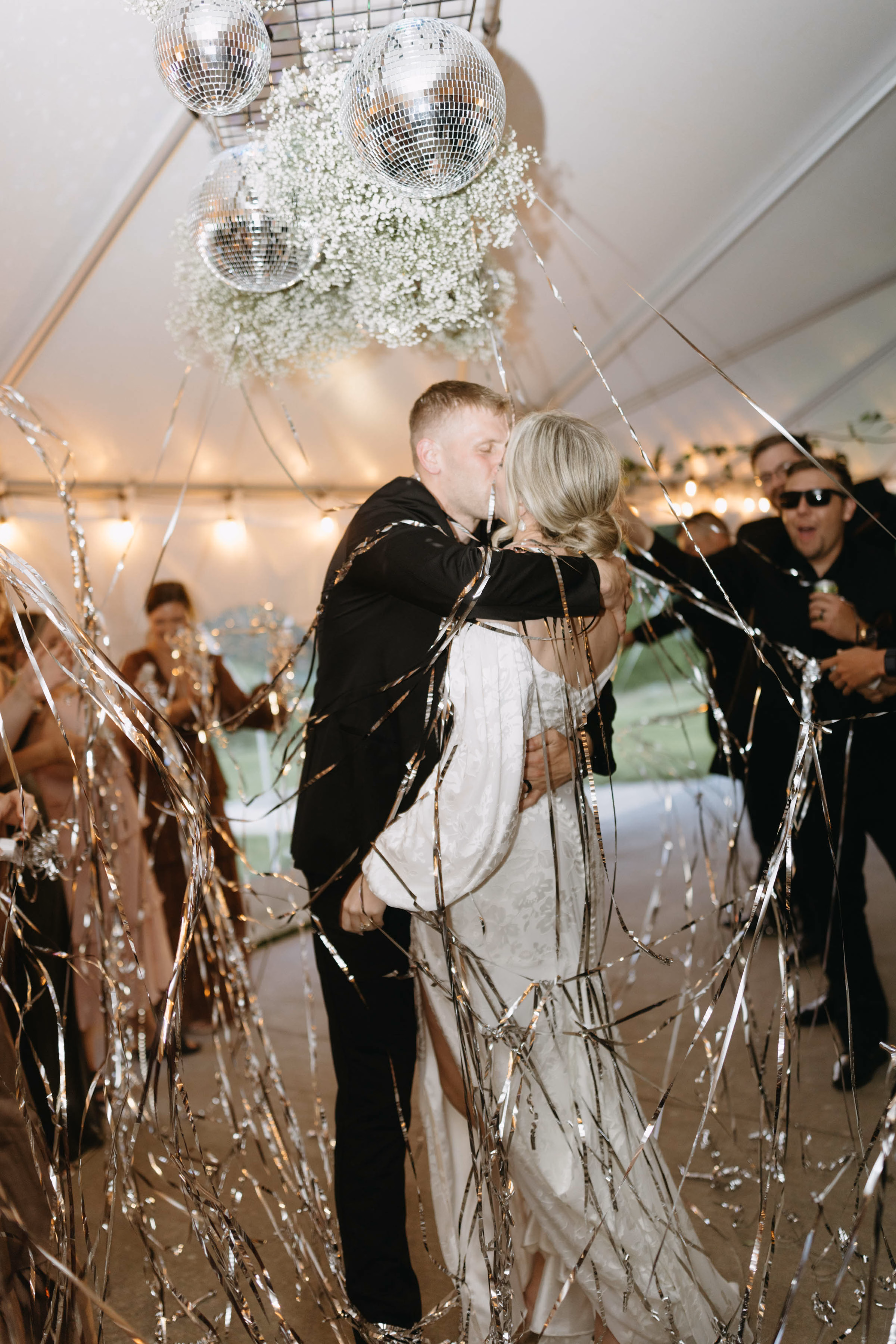 A bride and groom kiss on the dance floor as silver streamer cannons burst around them, with the high-energy reception flawlessly coordinated by Skies the Limit Events, a party planner in La Crosse, WI. The tented ceiling features her custom event design, showcasing an installation of shimmering disco balls and clouds of white baby's breath. Should we keep uplo