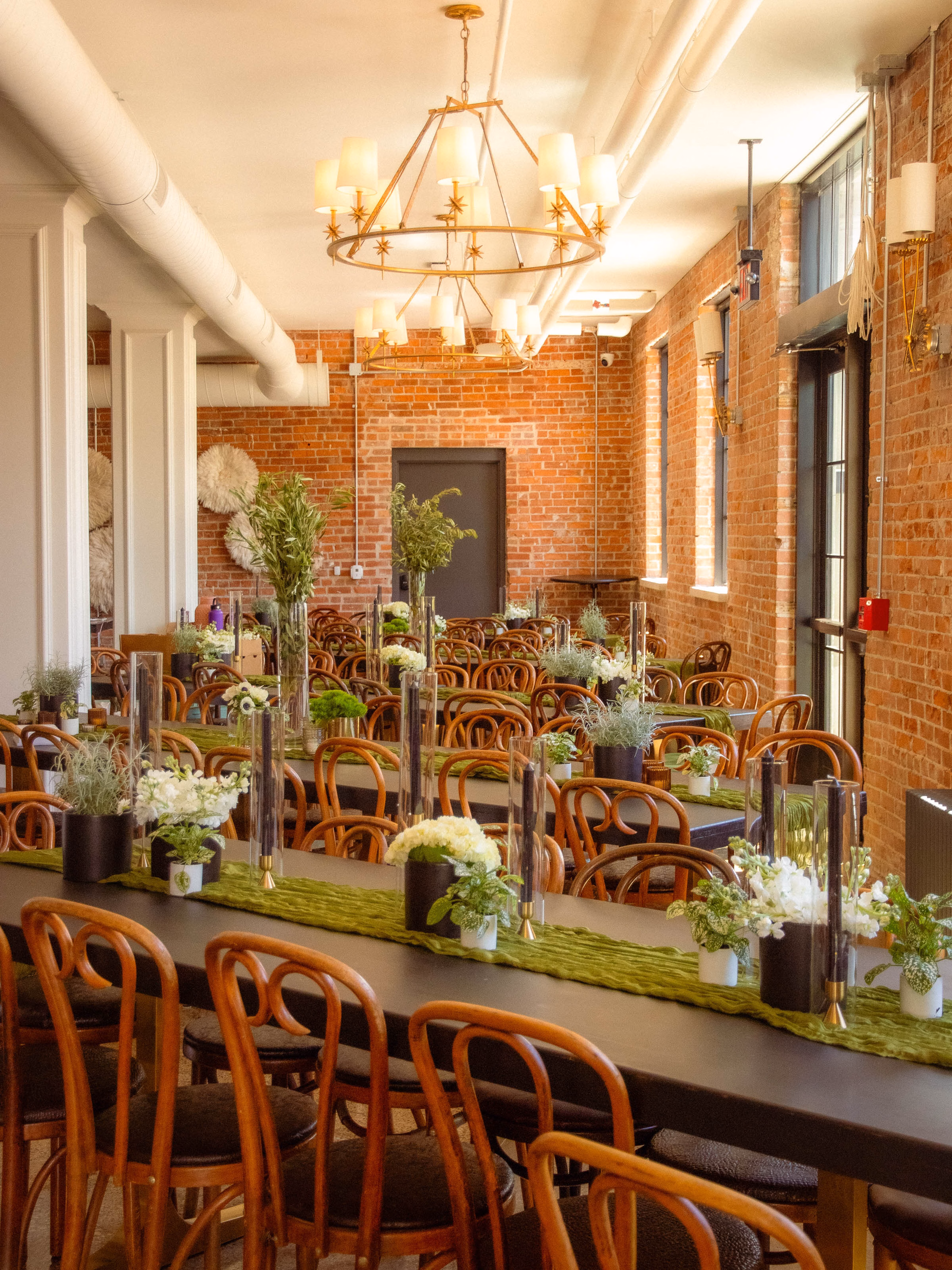 An industrial-chic reception room with exposed brick walls, beautifully styled and planned by Skies the Limit Events, a wedding and party planner in La Crosse, WI. The long banquet tables feature her custom design work, including vibrant green moss runners, crisp white floral centerpieces, tall glass cylinder candles, and classic wooden bentwood chairs under large statement chandeliers.