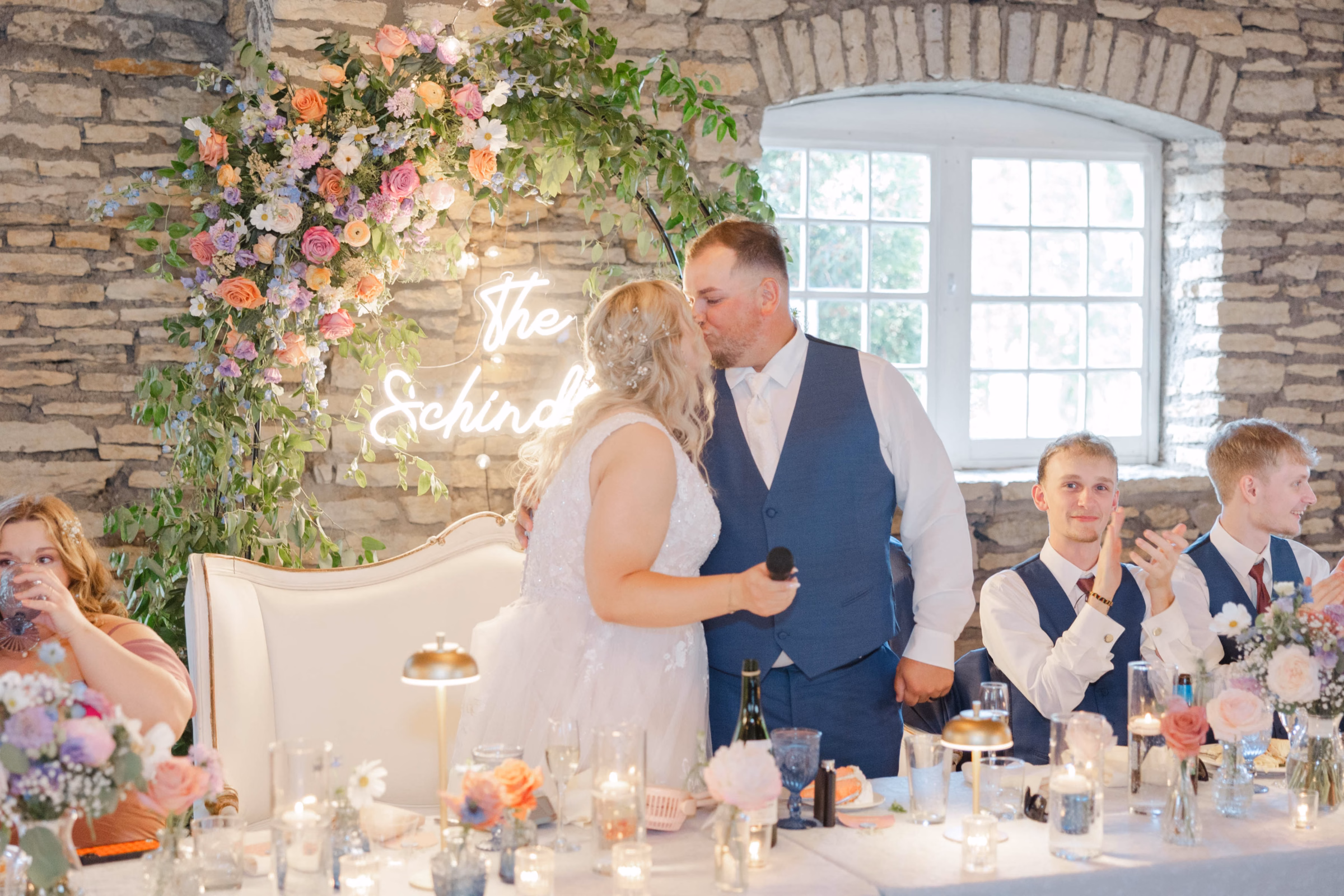 A bride and groom share a kiss at their head table in a rustic stone wall venue, beautifully planned and designed by Skies the Limit Events, a party planner in La Crosse, WI. The setup features a custom neon name sign framed by a vibrant, asymmetric floral arch with pastel and orange roses, matching small table centerpieces, and glowing candlelight.