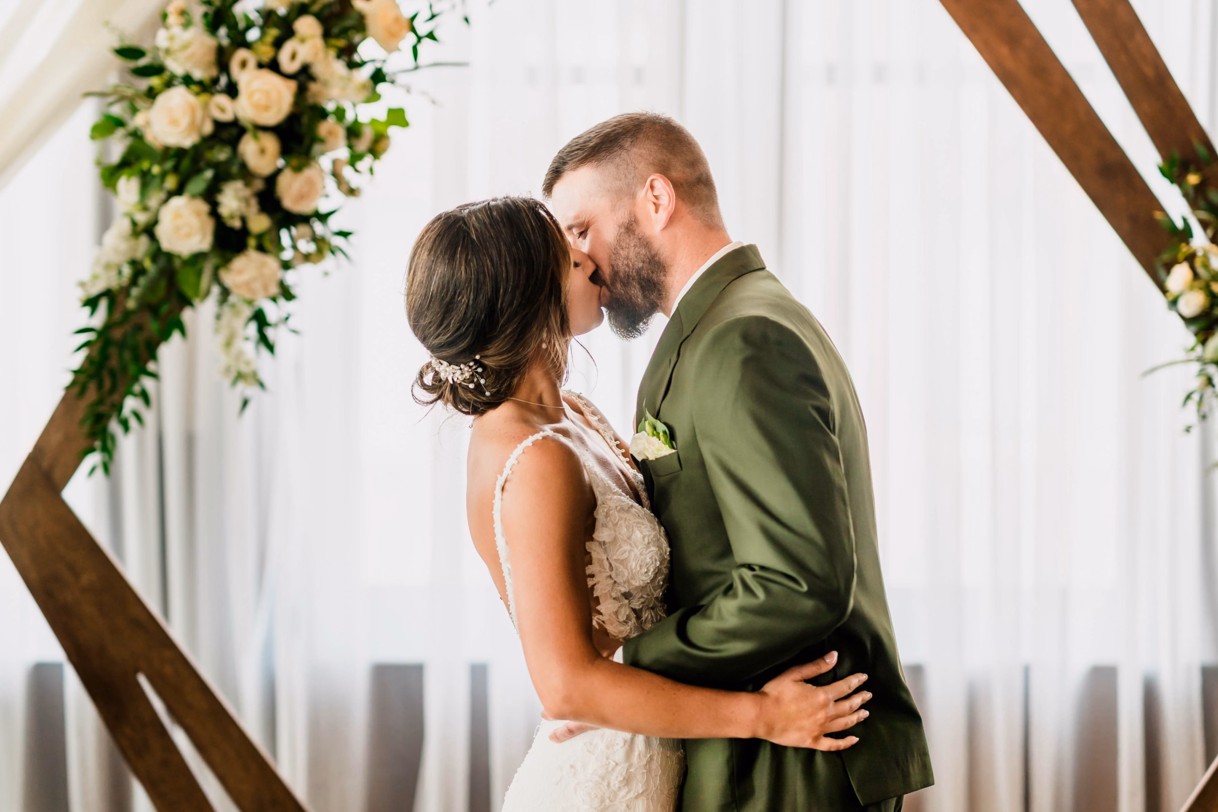 A bride and a groom in an olive green suit share a kiss under a geometric wooden arch, with the wedding day flawlessly organized by Skies the Limit Events, a wedding planner and party planner in La Crosse, WI. The modern indoor altar setup is accented by her custom floral arrangements featuring white roses and crisp greenery.