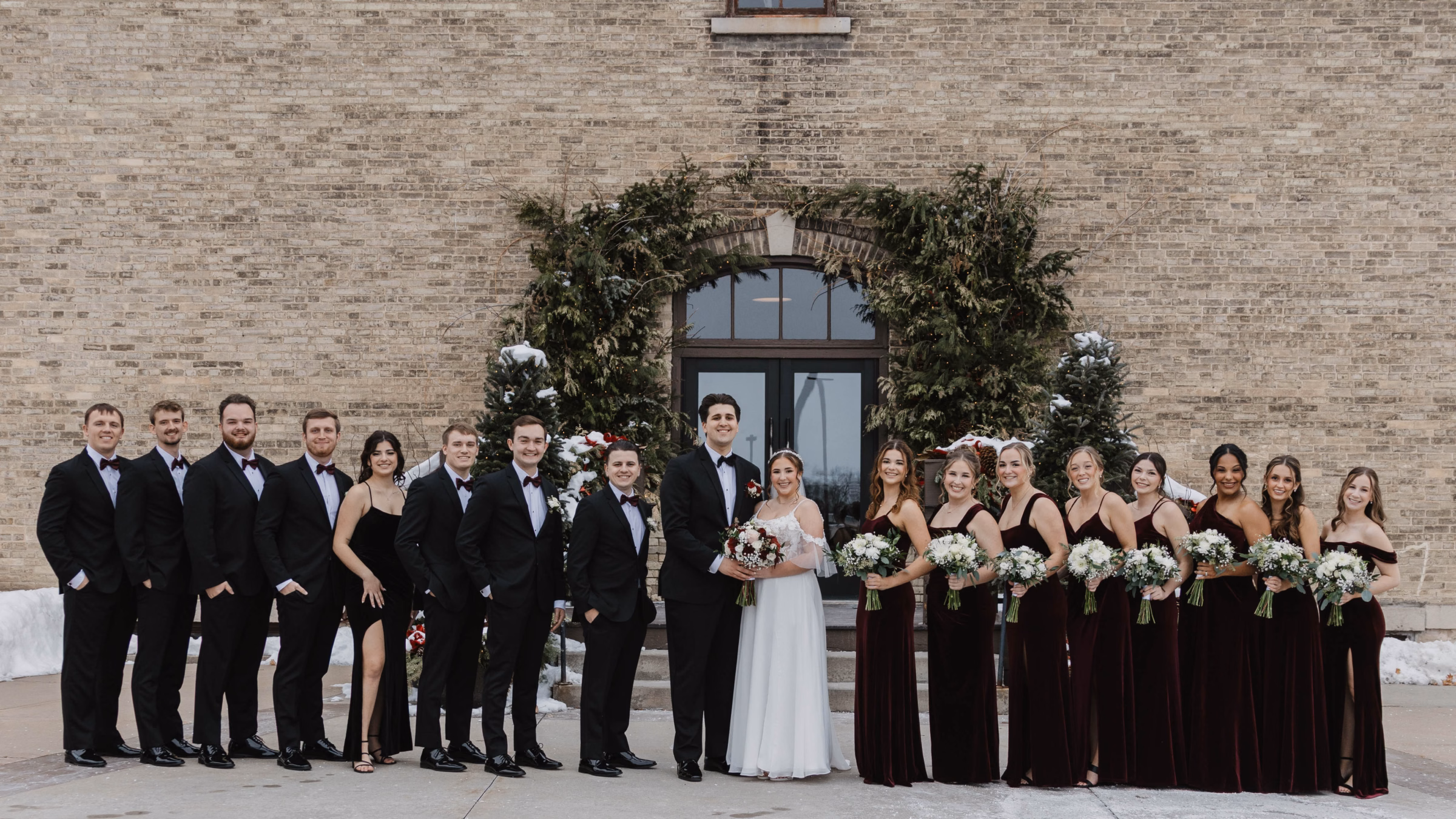 A large winter wedding party smiles outside a historic cream brick building framed with a lush evergreen arch, highlighting the bridesmaids in rich burgundy velvet gowns and a classic black-tie groom, captured by Abigail Peters Photography.