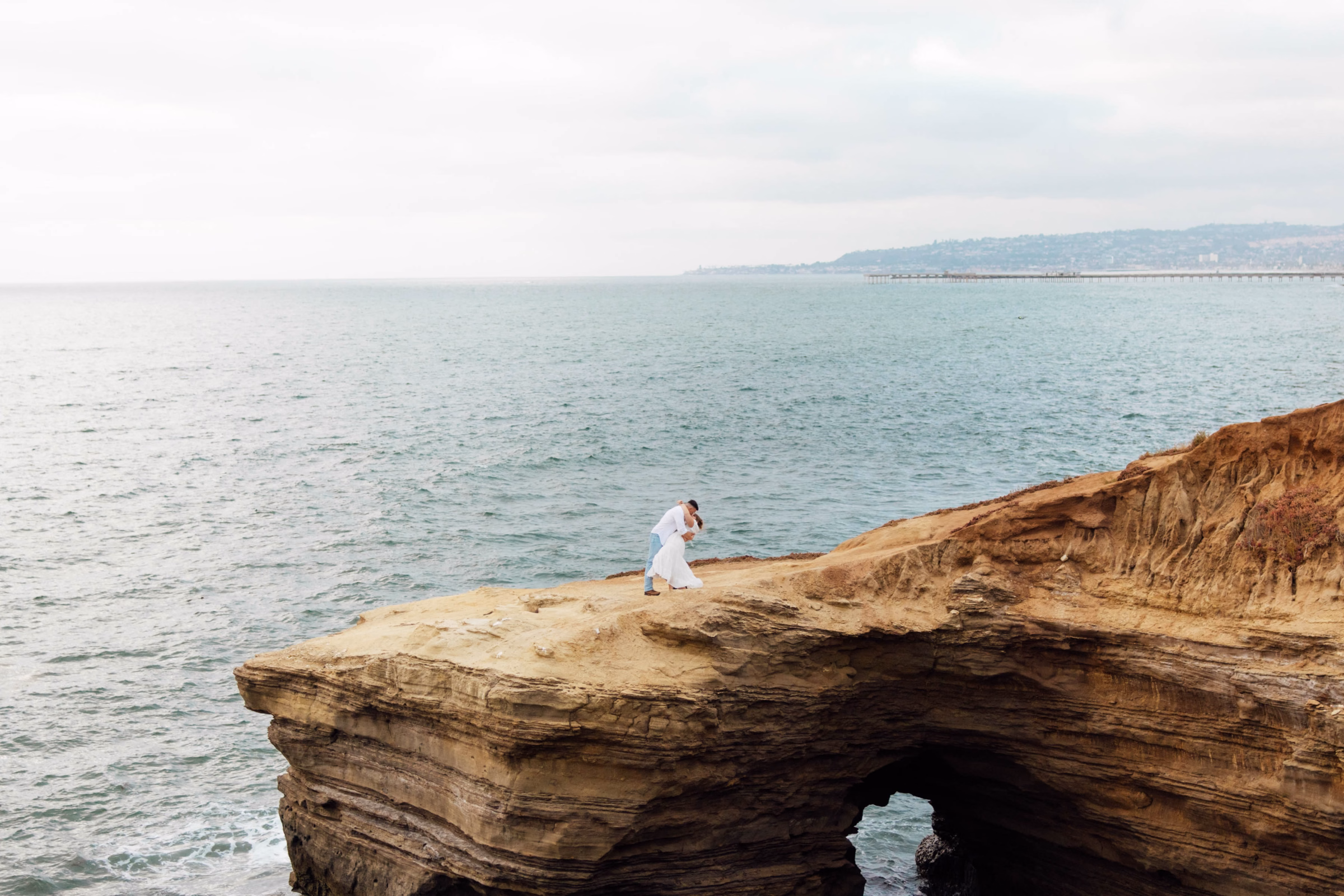 An expansive wide shot of a couple kissing on a high, rugged ocean cliffside above a sea cave arch, capturing the vast blue water and a distant pier, photographed by Abigail Peters Photography.