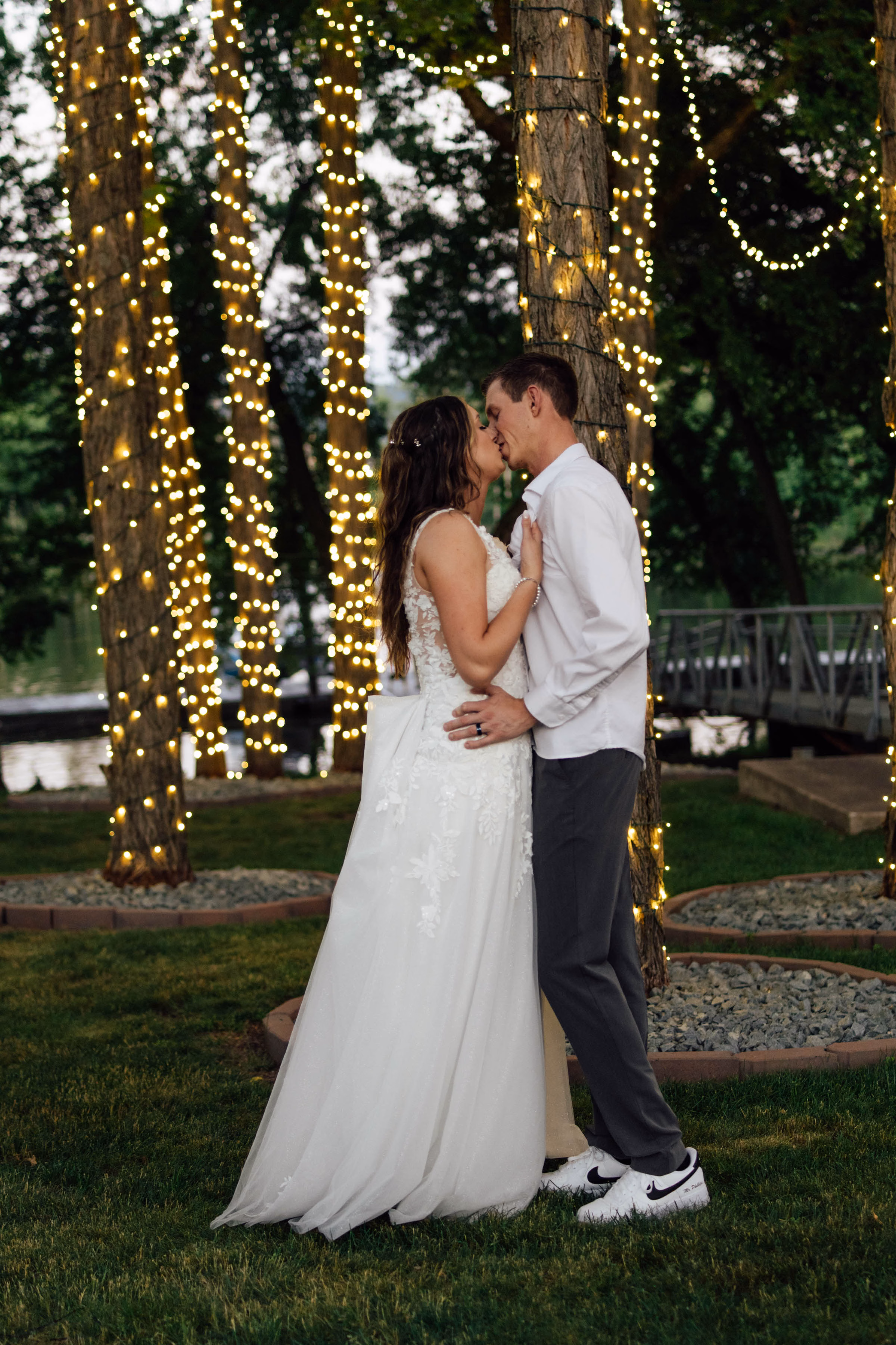 A bride in a lace-detailed a-line gown and a groom in a casual white shirt kiss outdoors under trees wrapped in glowing fairy lights, captured by Abigail Peters Photography.