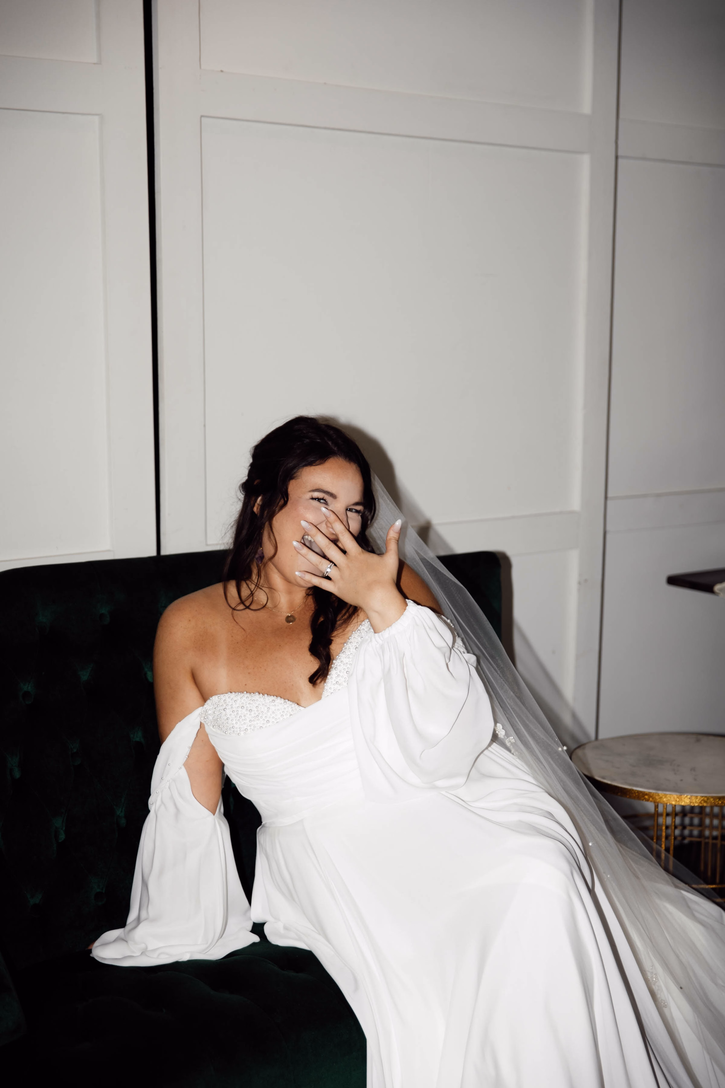 A joyful bride laughs candidly while sitting on a dark green velvet couch, showing off her engagement ring and an off-the-shoulder gown with balloon sleeves, captured by Abigail Peters Photography.