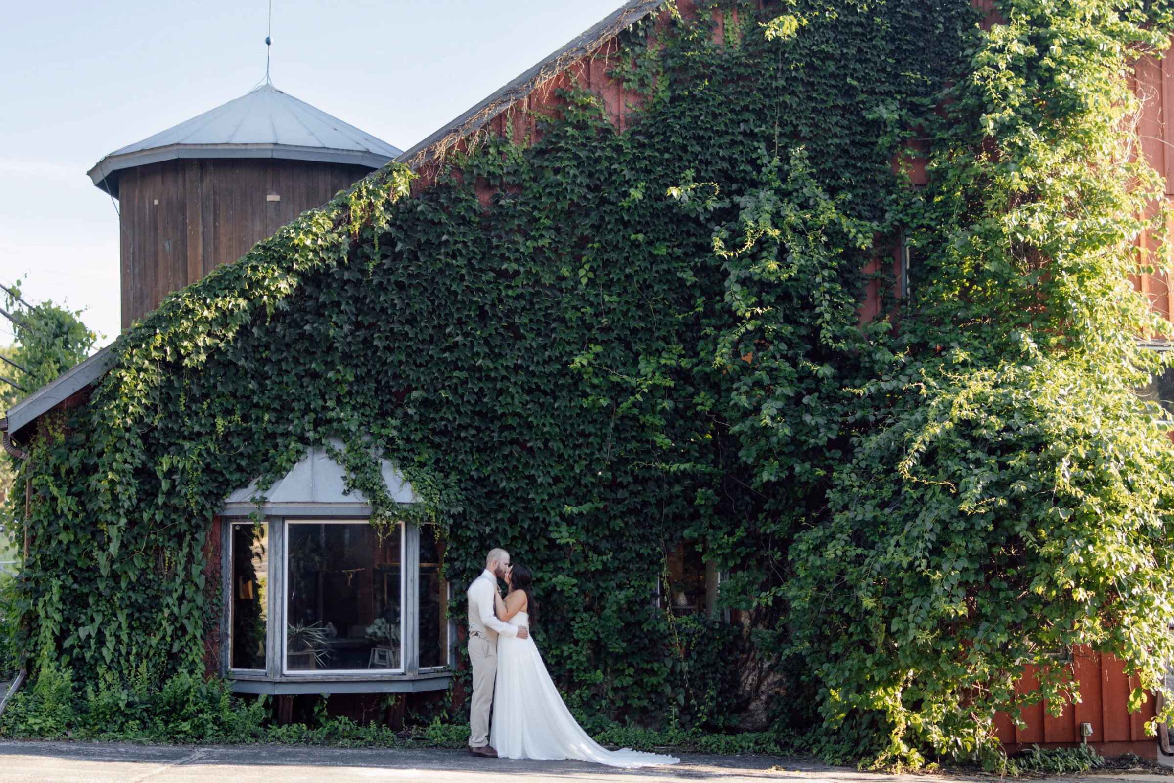 A bride and groom share a romantic embrace outside a rustic red barn covered in a massive wall of lush green ivy, featuring a wooden silo and a large bay window, captured by Abigail Peters Photography.