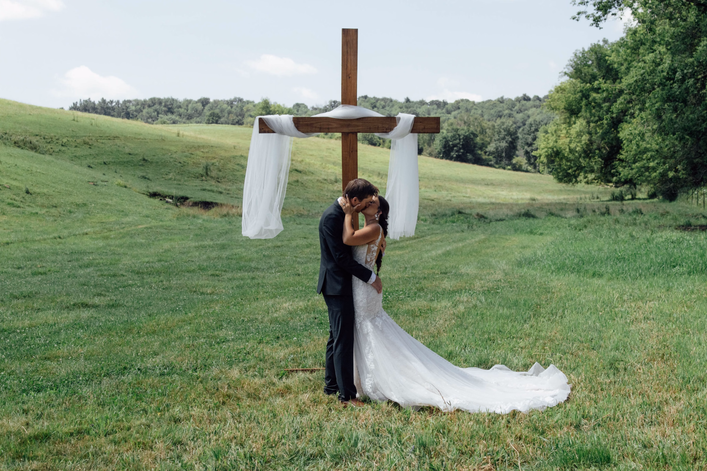 A bride and groom kiss during an outdoor countryside ceremony in a rolling green field, standing in front of a rustic wooden cross draped in flowing white tulle, captured by Abigail Peters Photography.
