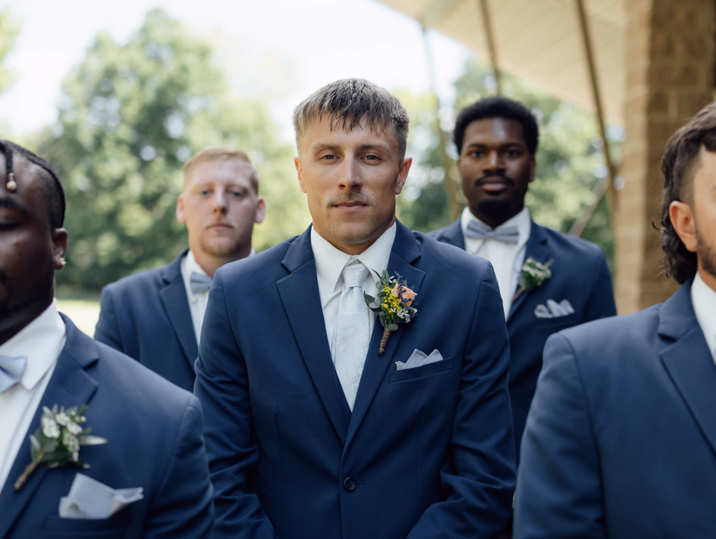 A groomsmen lineup features a groom in a classic navy blue wedding suit with a subtle floral patterned white tie and a bright wildflower boutonniere, captured by Abigail Peters Photography.