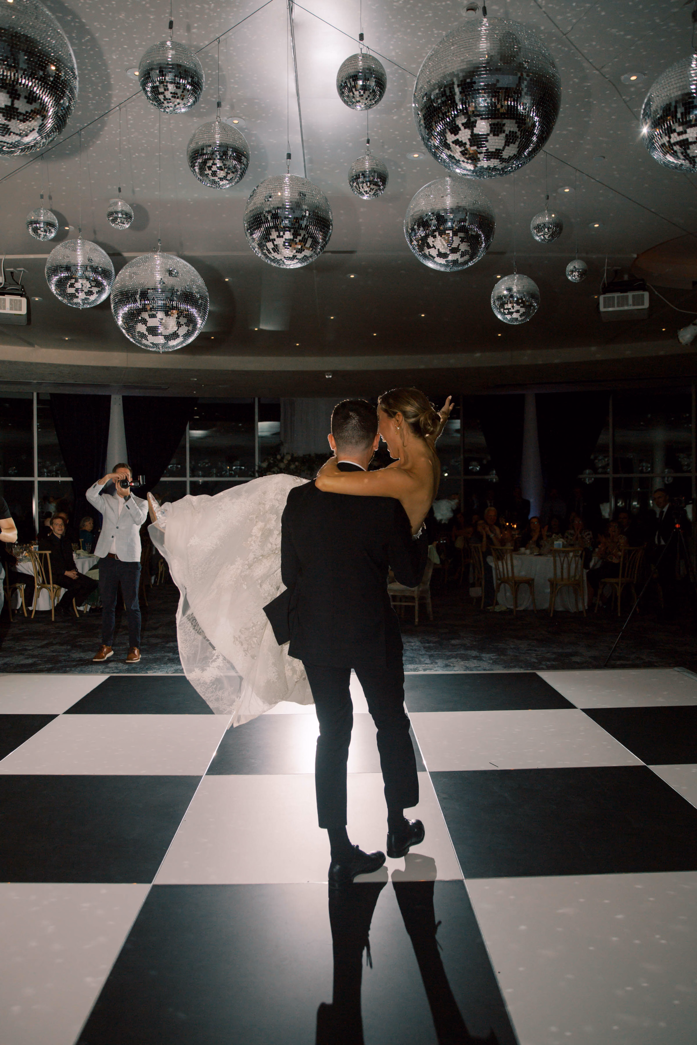 An Event Essentials black and white checkered dance floor installed for a wedding reception, featuring a groom carrying his bride under an overhead cluster of multiple hanging disco balls.