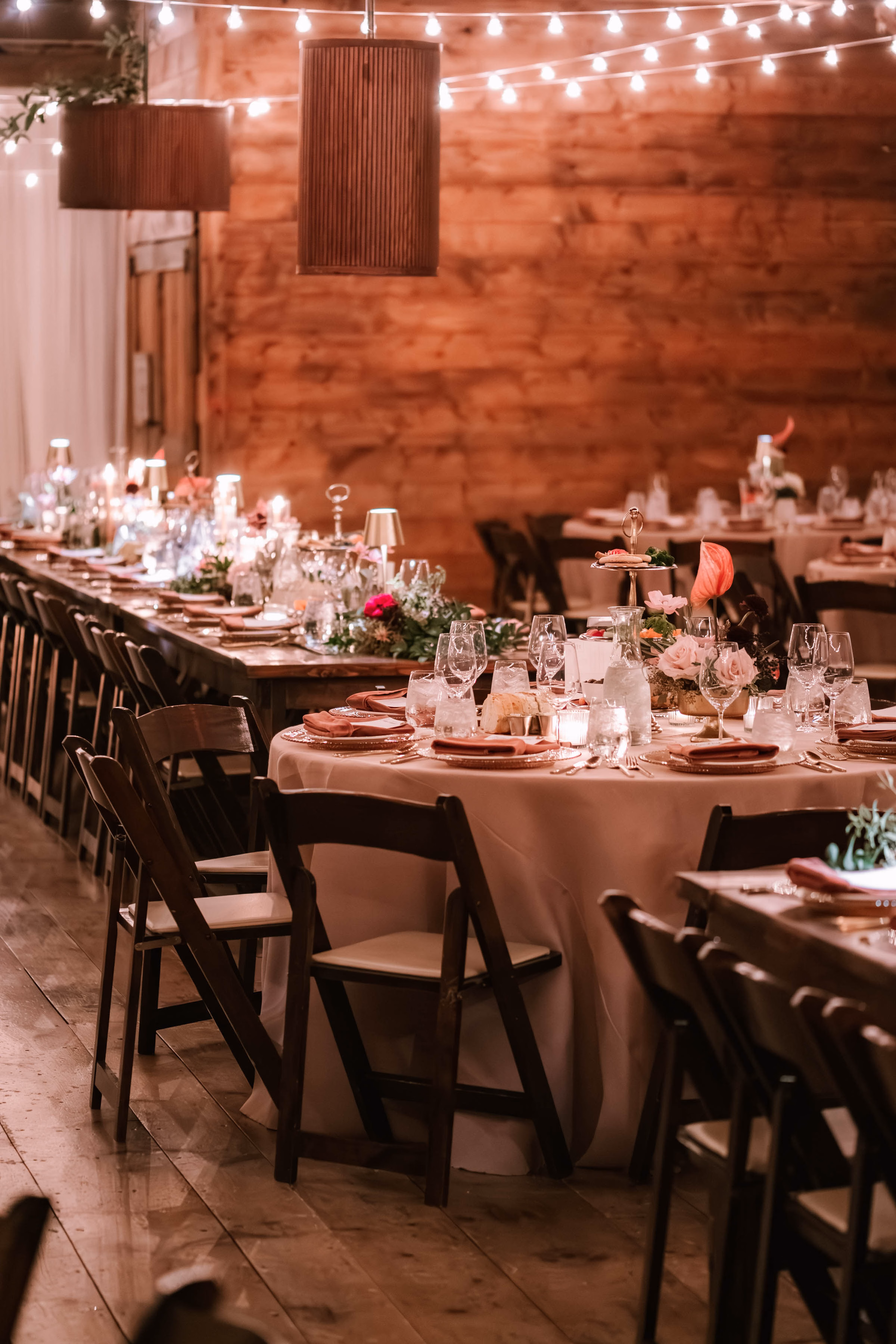An Event Essentials barn wedding reception setup featuring wooden farm tables mixed with round tables covered in blush linens, paired with dark wood folding chairs with white cushions under warm string lighting.