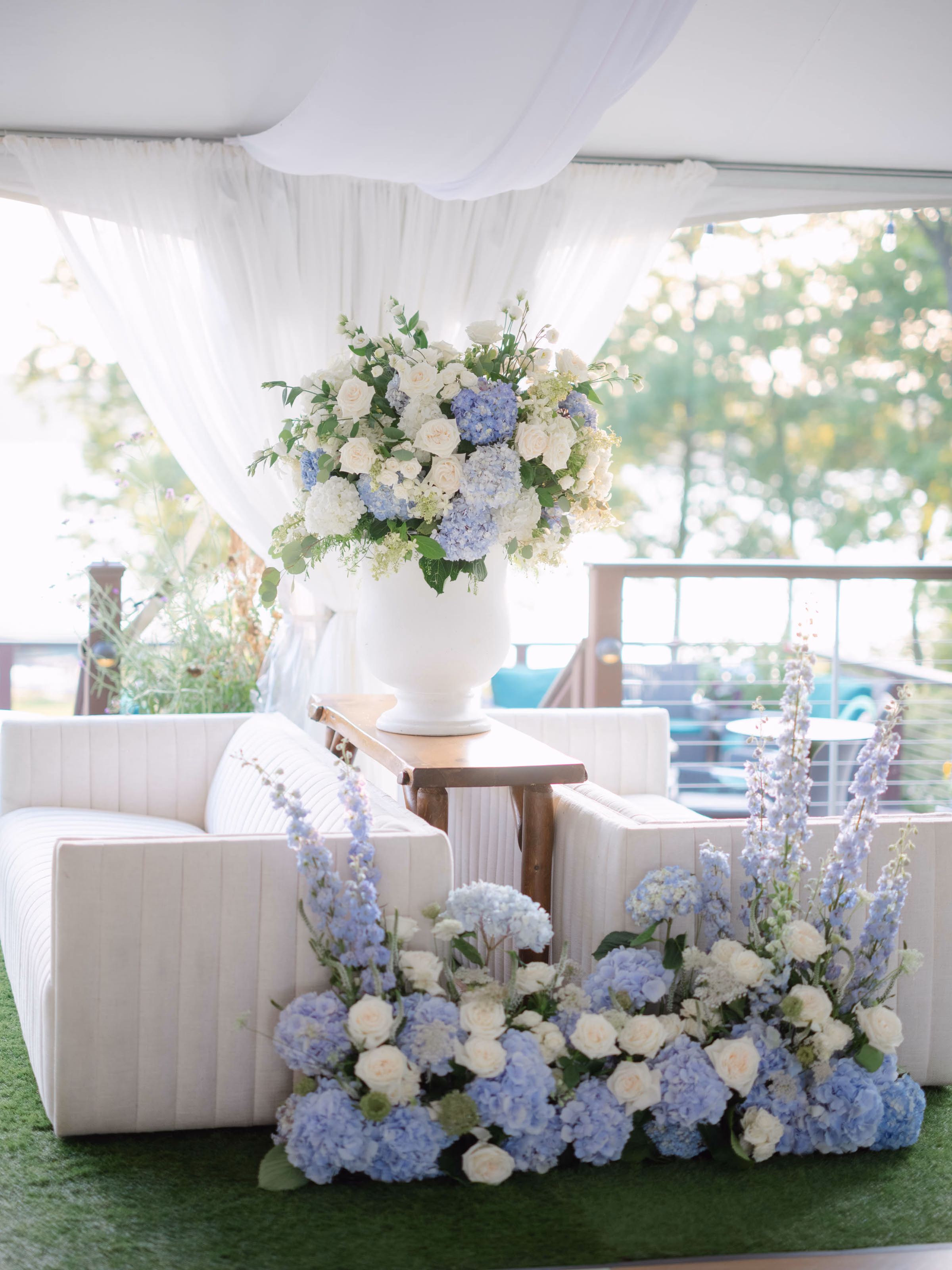 An Event Essentials outdoor event lounge featuring two white channel-tufted sofas facing each other, divided by a wooden console table with a large floral arrangement and white fabric drapery in the background.