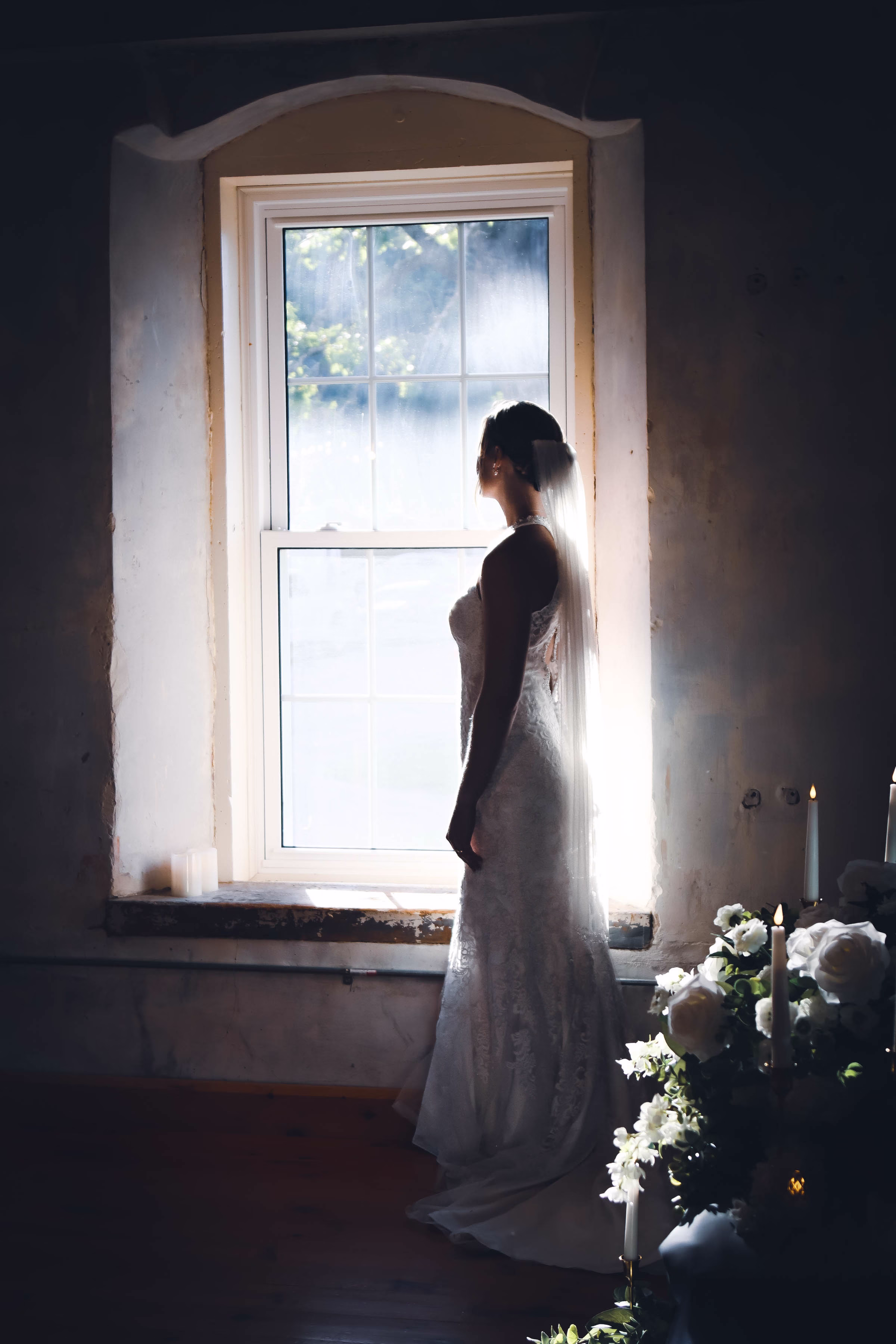 Silhouette of a bride in a veil looking out a sunlit window, candles and white florals in foreground at The Mill on Rush Creek