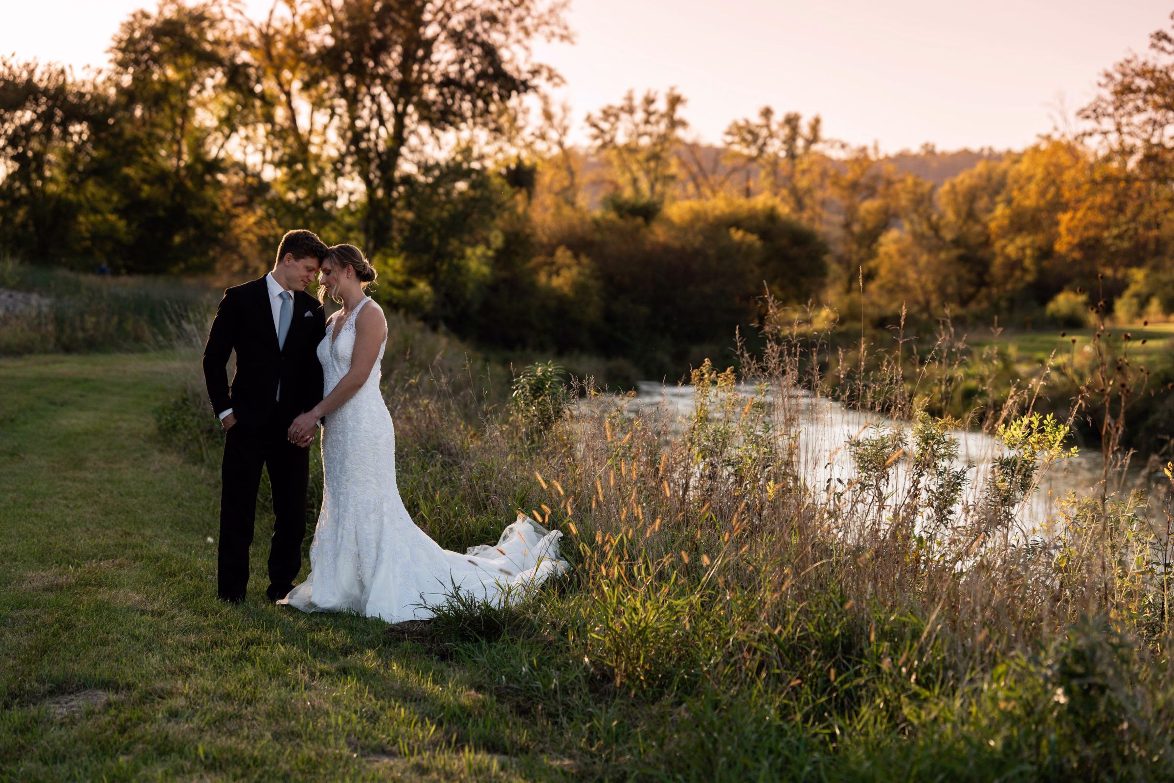 Bride and groom standing together at sunset along Rush Creek, surrounded by tall grass and golden fall trees