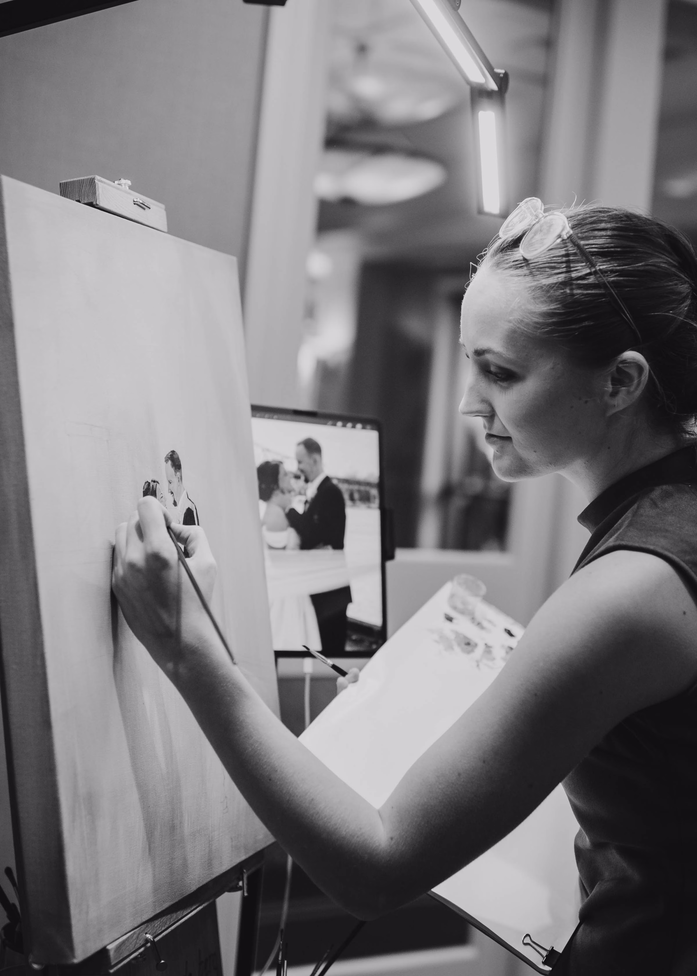 A black and white action photograph of live wedding painter Brenda Berg at work. She is captured from the side with glasses pushed up on her head, looking intently at her canvas as she paints a fine detail with a thin paintbrush. A professional easel light illuminates the canvas, and a digital reference screen displaying a photo of a bride and groom standing close together stands behind her.