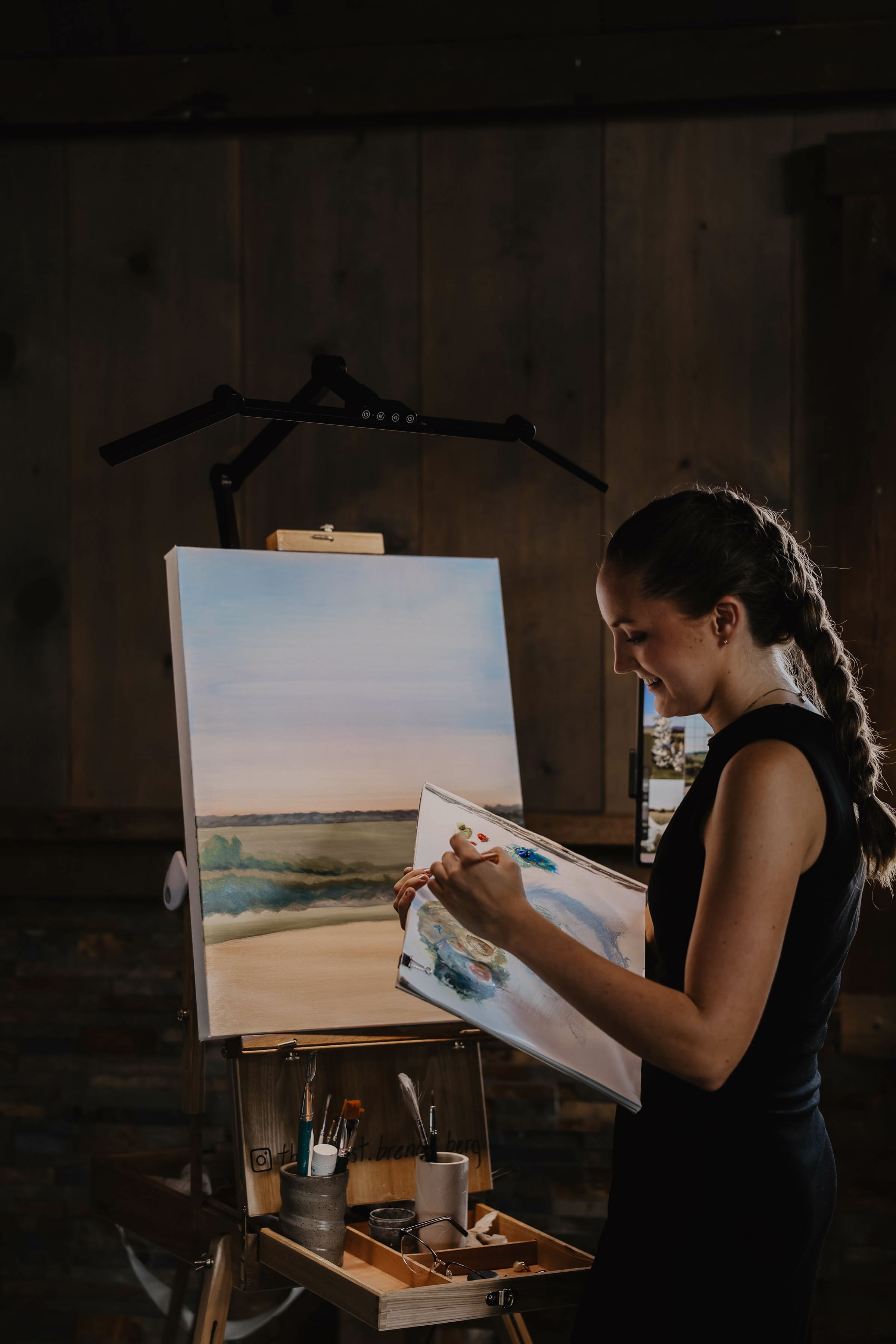 A warm, moody action photograph of live wedding painter Brenda Berg smiling as she works at her wooden easel indoors. Her hair is in a long braid and she wears a black sleeveless dress while holding her palette and mixing paint with a brush. An overhead professional light illuminates her canvas, which depicts a scenic landscape painting of a field and shoreline under a soft, pale sky.
