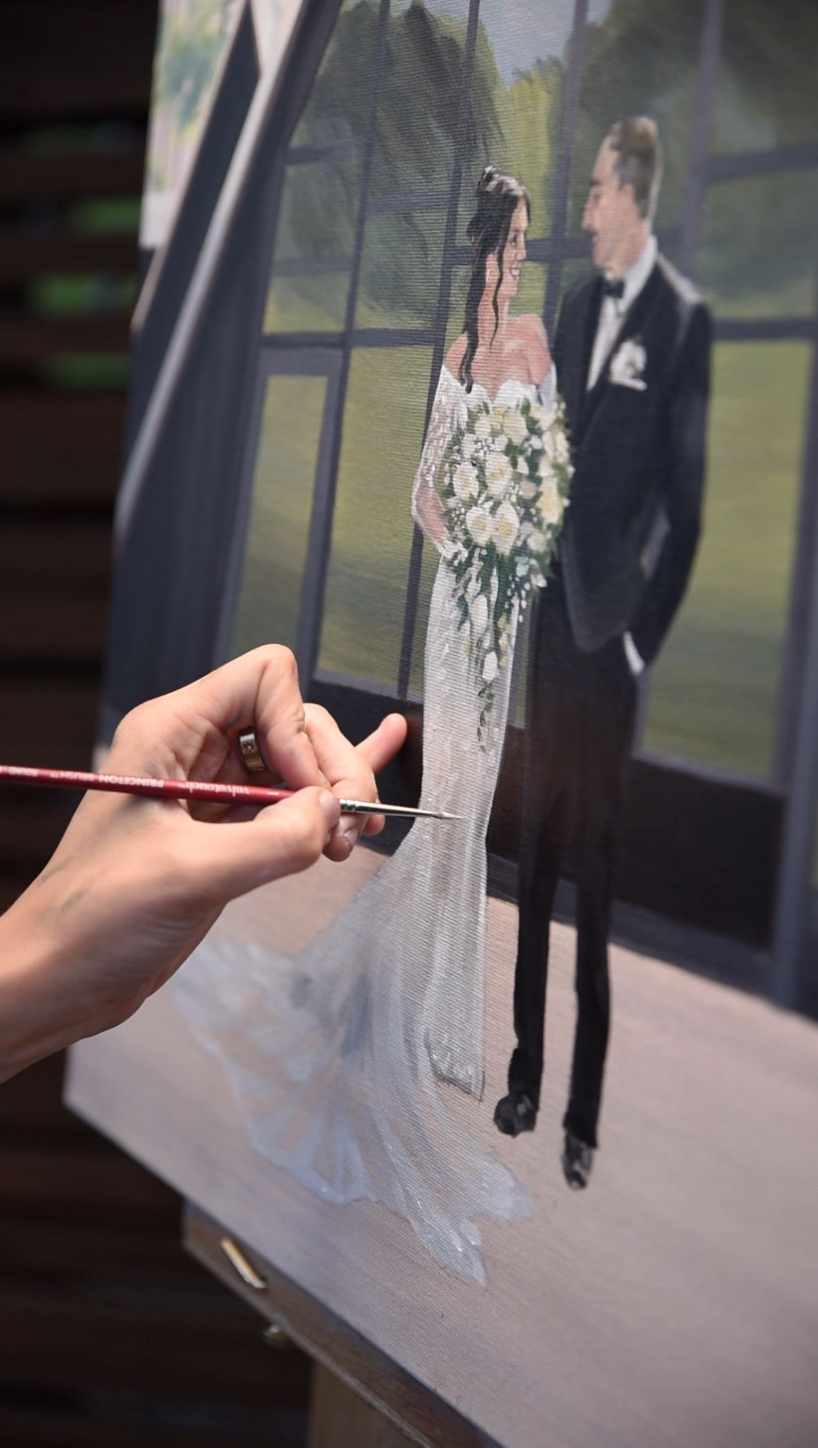An extreme close-up, angled action photograph highlighting live wedding painter Brenda Berg’s hand holding a thin brush as she paints fine details onto a canvas. The painting depicts a bride in an elegant off-the-shoulder lace gown holding a large white rose bouquet, smiling at her groom who wears a classic black tuxedo, set against a modern dark-framed window backdrop.