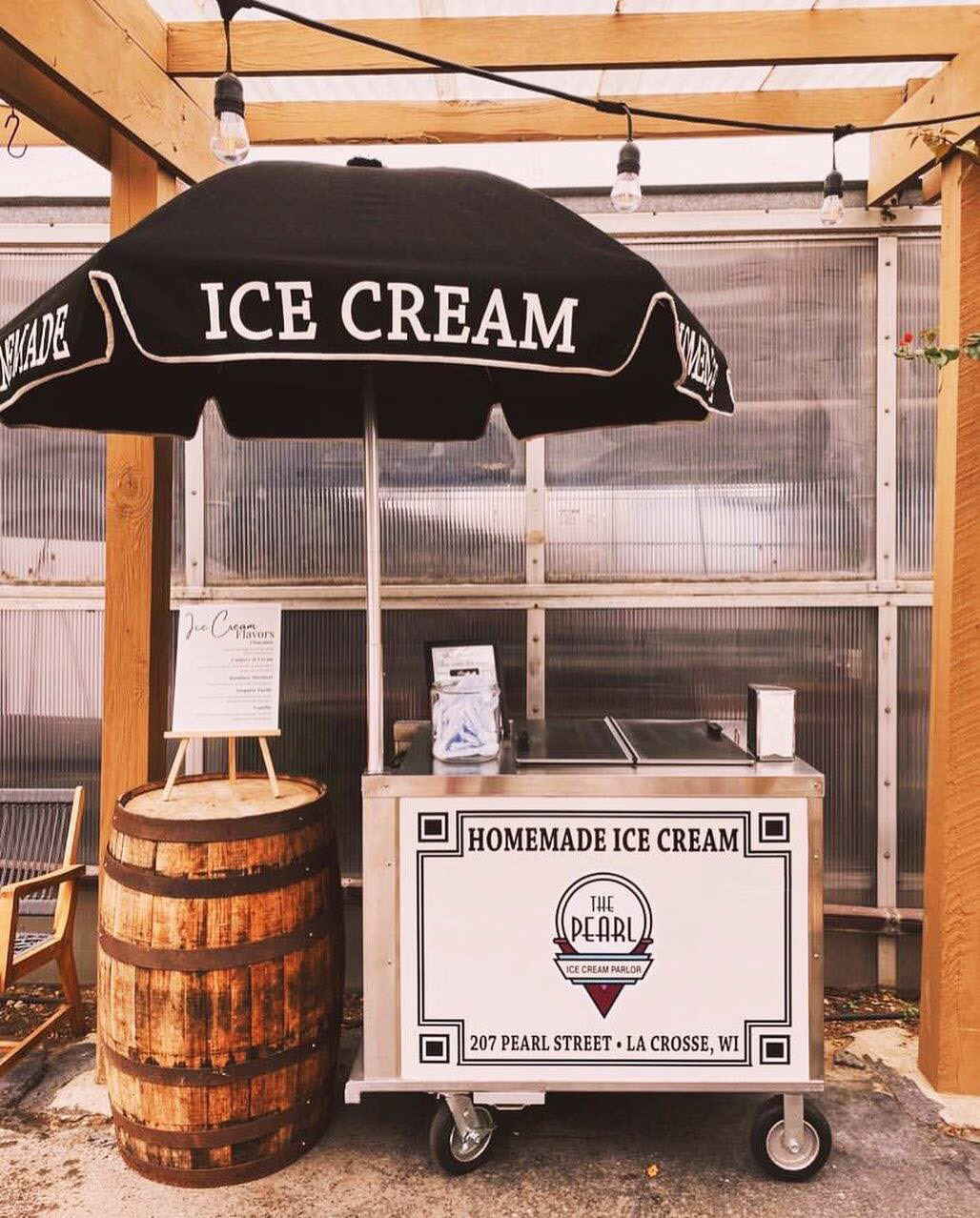 An Ice cream cart branded with The Pearl Ice Cream Parlor logo and an umbrella that says Homemade Ice Cream La Crosse, WI, next to a barrel at a rustic wedding venue.