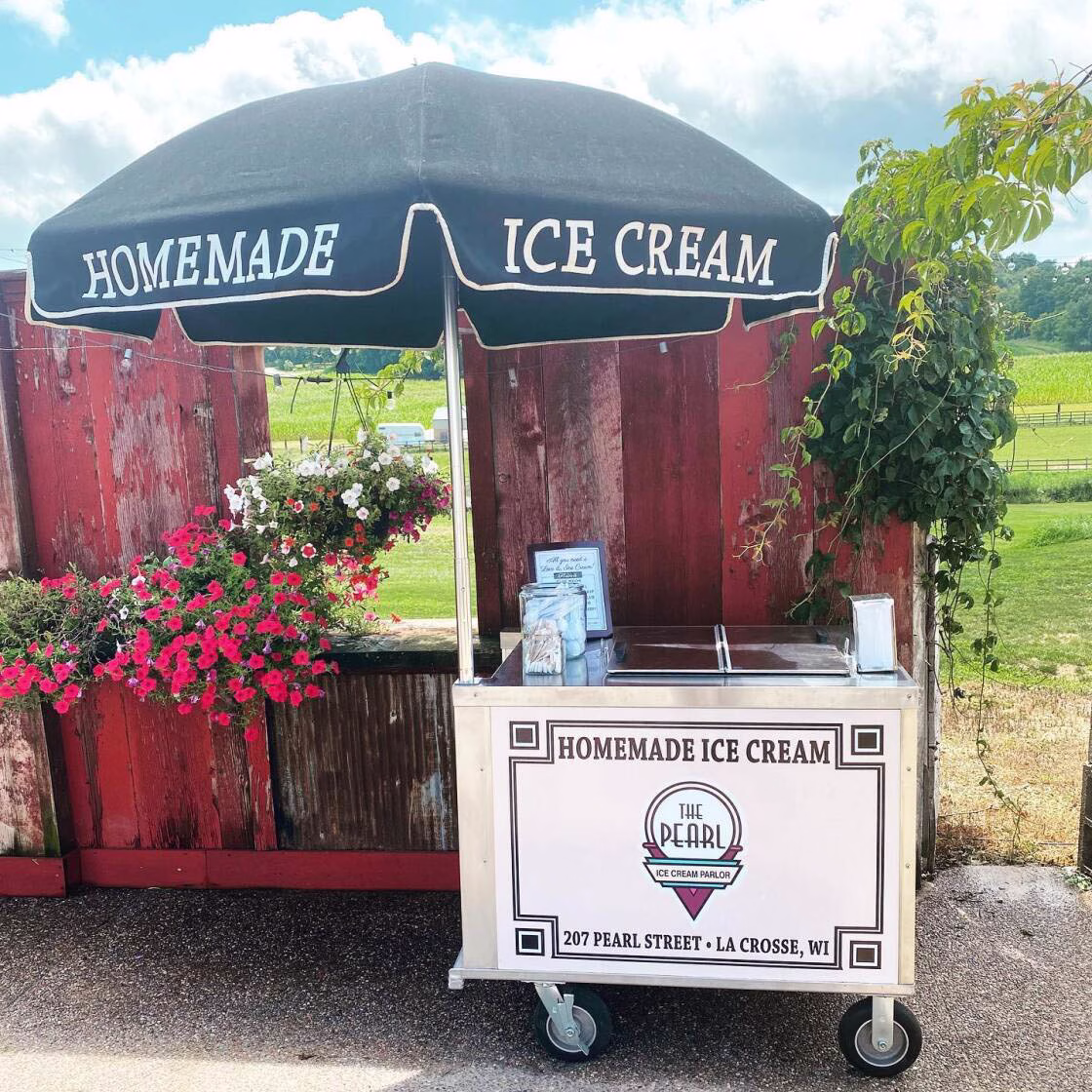 An Ice cream cart branded with The Pearl Ice Cream Parlor logo and an umbrella that says Homemade Ice Cream La Crosse, WI, in front of a red wall