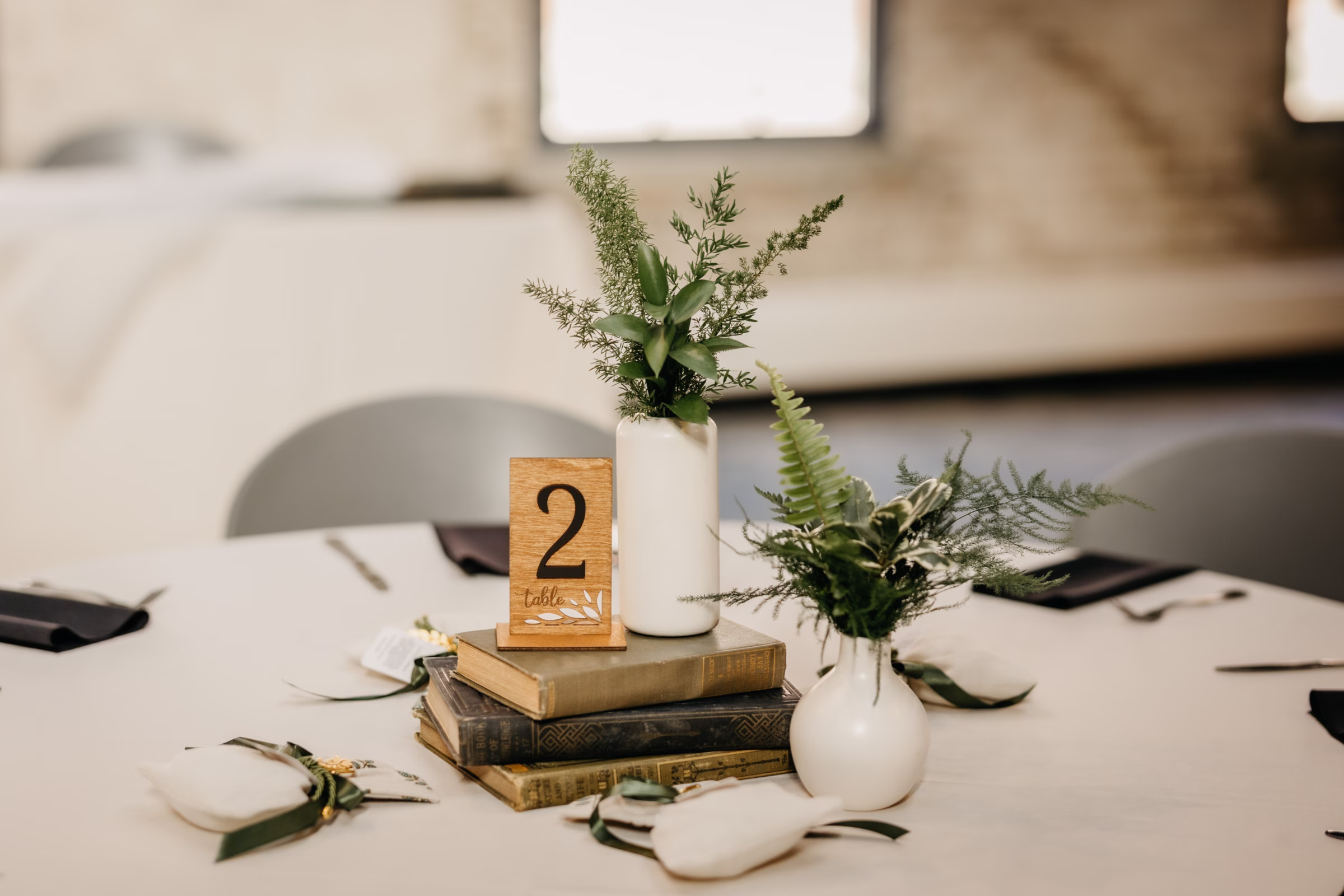 a detail photo showing a wedding reception table centerpiece made from vintage stacked books. The table number is made of wood, and the centerpiece also includes vases of ferns and greenery. Greek wedding favors surround the center piece