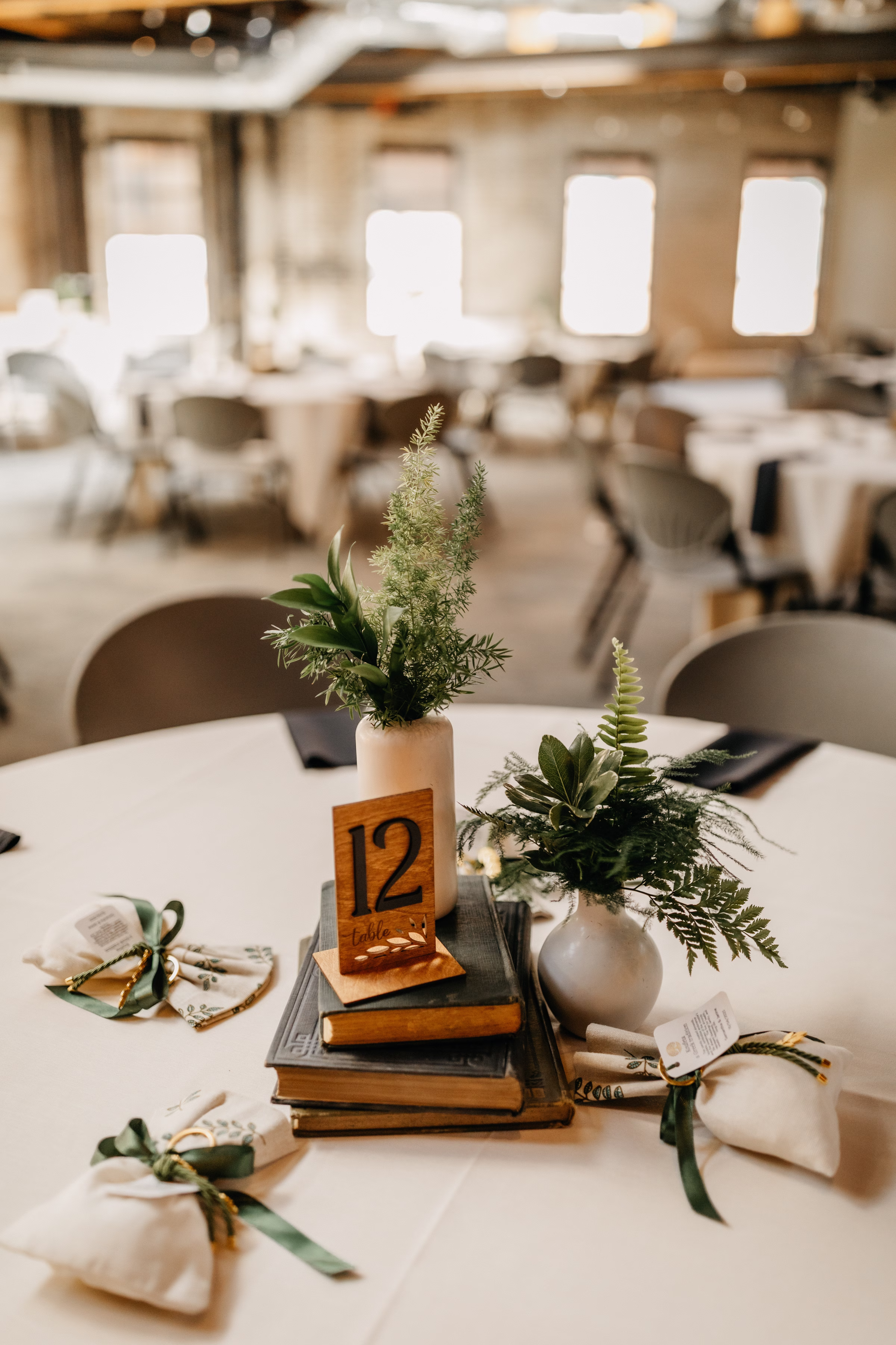 a detail photo showing a wedding reception table centerpiece made from vintage stacked books. The table number is made of wood, and the centerpiece also includes vases of ferns and greenery. Greek wedding favors surround the center piece