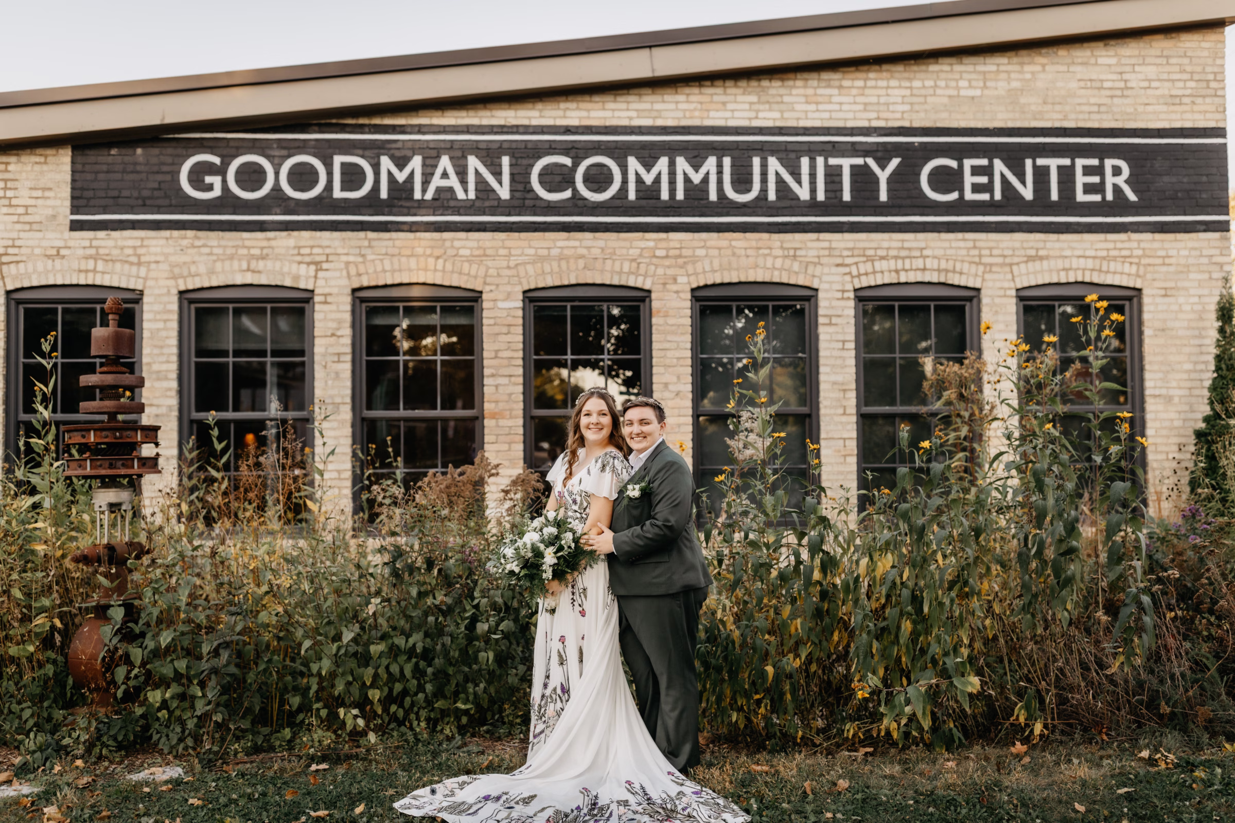 the brides pose in front of Goodman Community Center, framed by natural Wisconsin flowers and foliage during a portrait session at their organic & authentic wedding