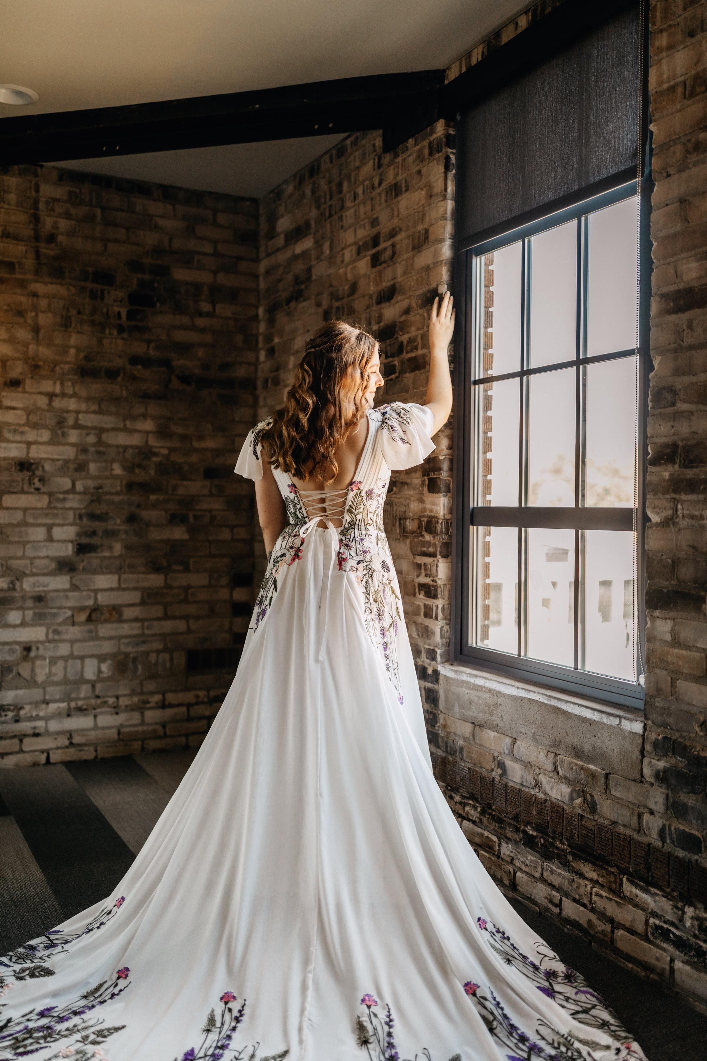 a bride stands looking outside a window, showing off the back of her wedding dress with ribbon corset and colorful floral and vine embroidery