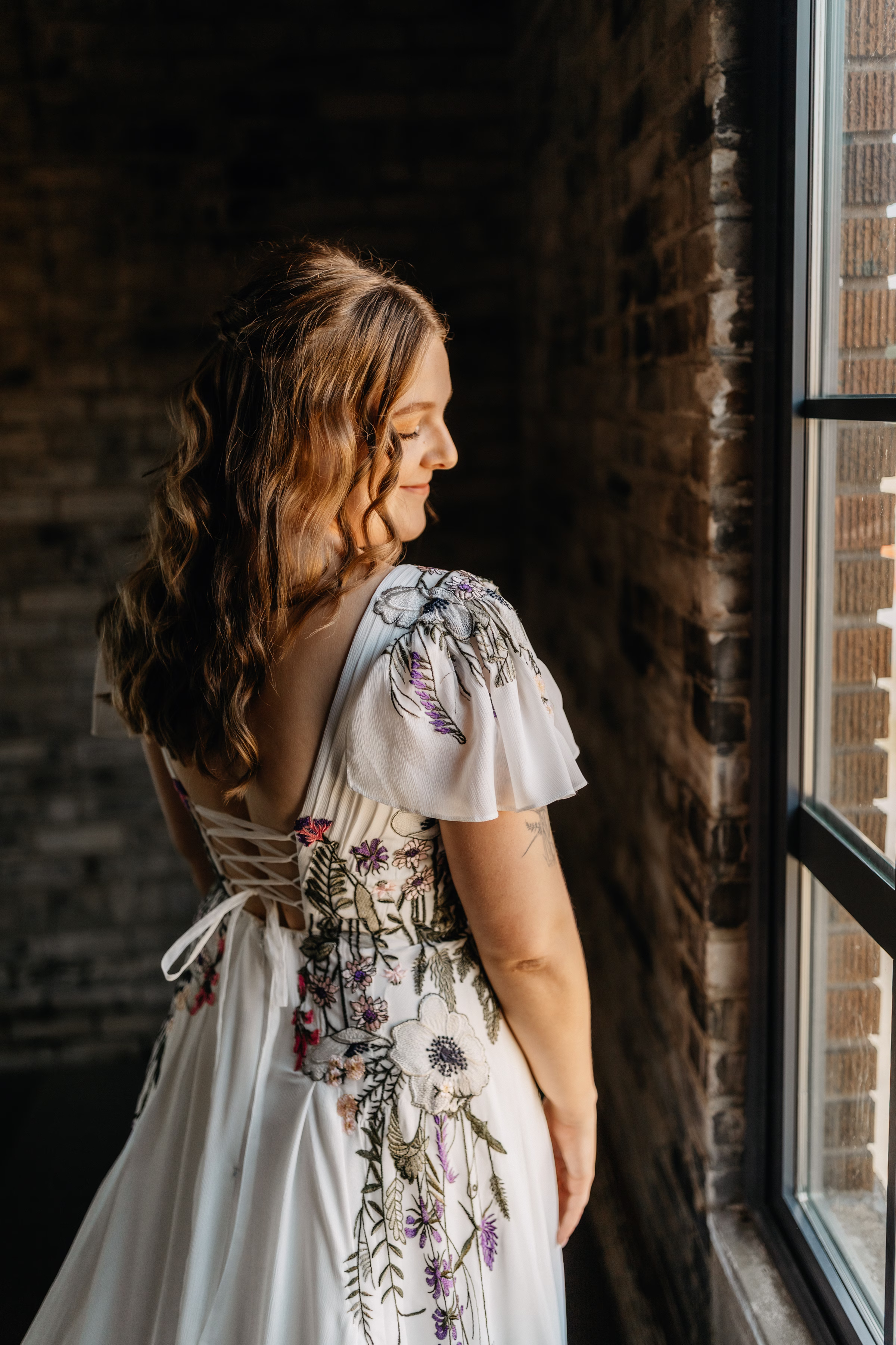 a bride shows off her hair and the back of her wedding dress featuring airy sleeves, a tied corset, and colorful embroidery