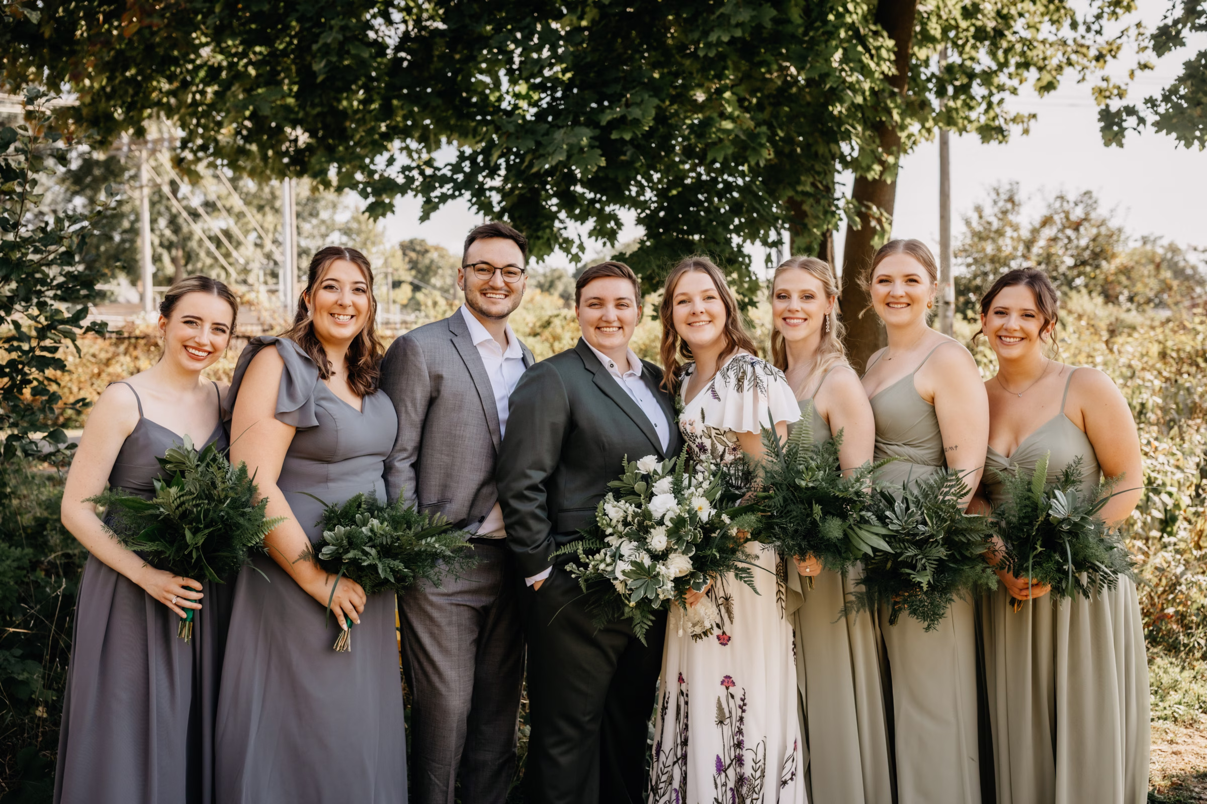Sam and Janine pose with their wedding party, dressed in pale sage green and gray. Sam holds her bridal bouquet, and the wedding party holds greenery bouquets
