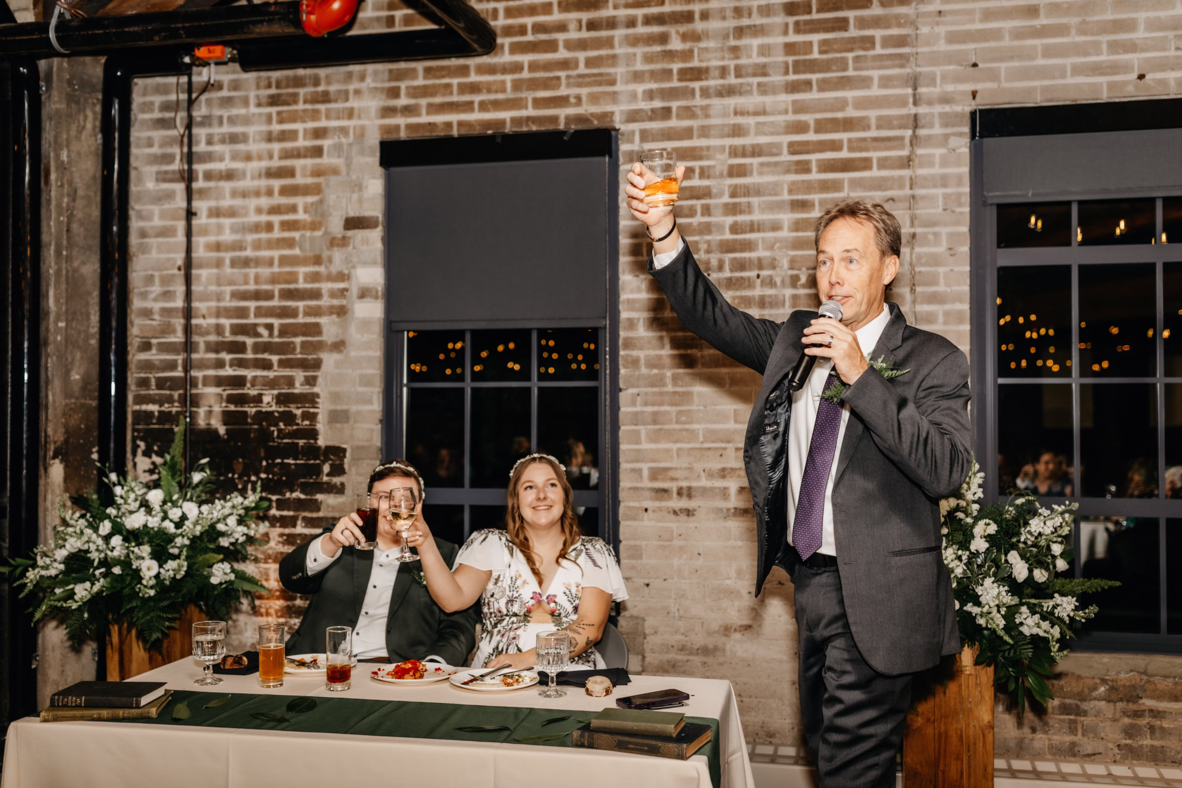 the brides smile at their sweetheart table while a guest starts a toast