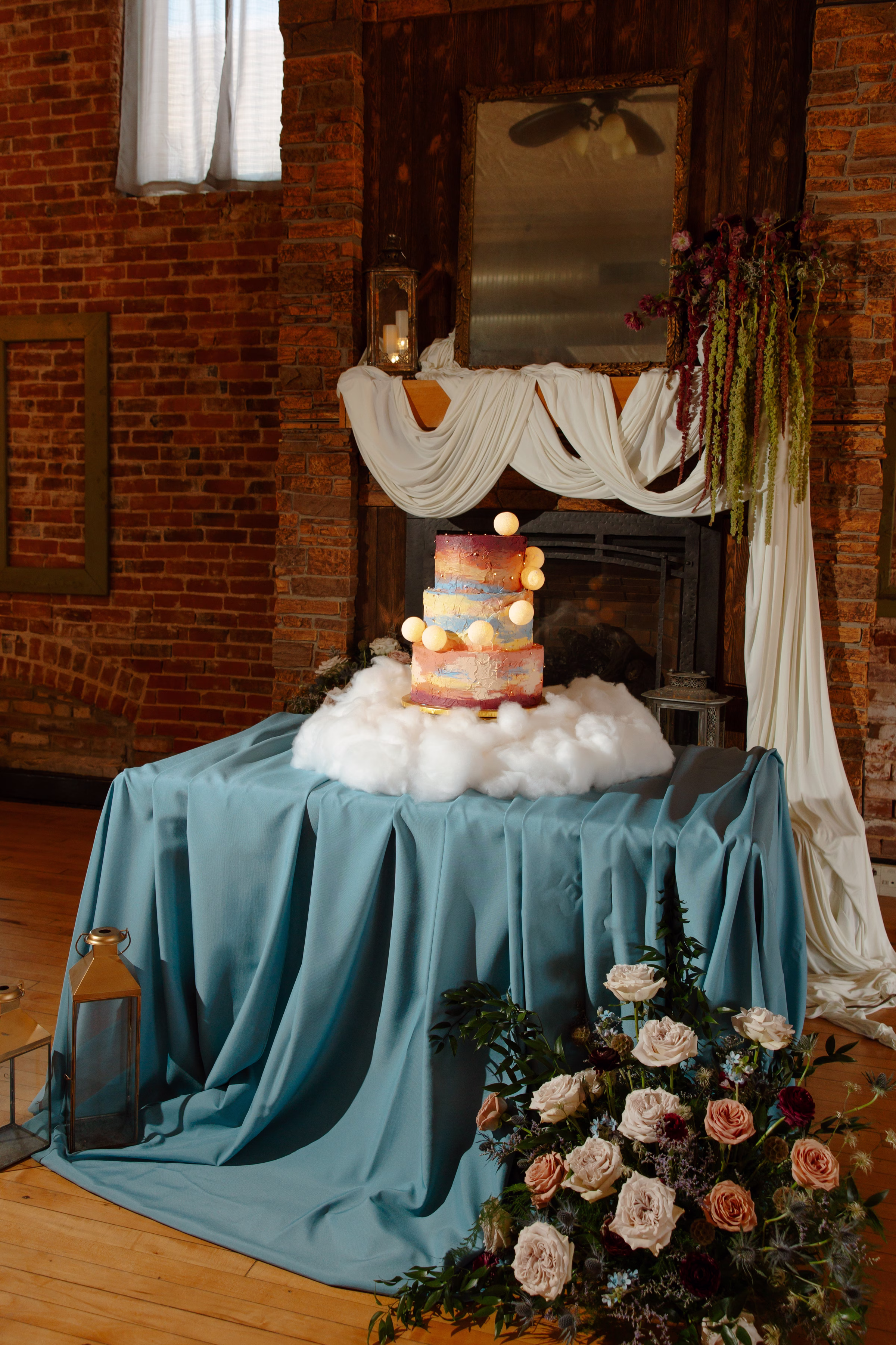 a celestial summer sunset wedding cake display by Cowboy David's Bakery with painted frosting and playful moons. The cake sits on faux clouds on a table with a blue tablecloth in front of the fireplace at B's Place Event Venue in West Salem, Wisconsin. Florals from Bloom & Knot LLC and lanterns from Event Essential are also present