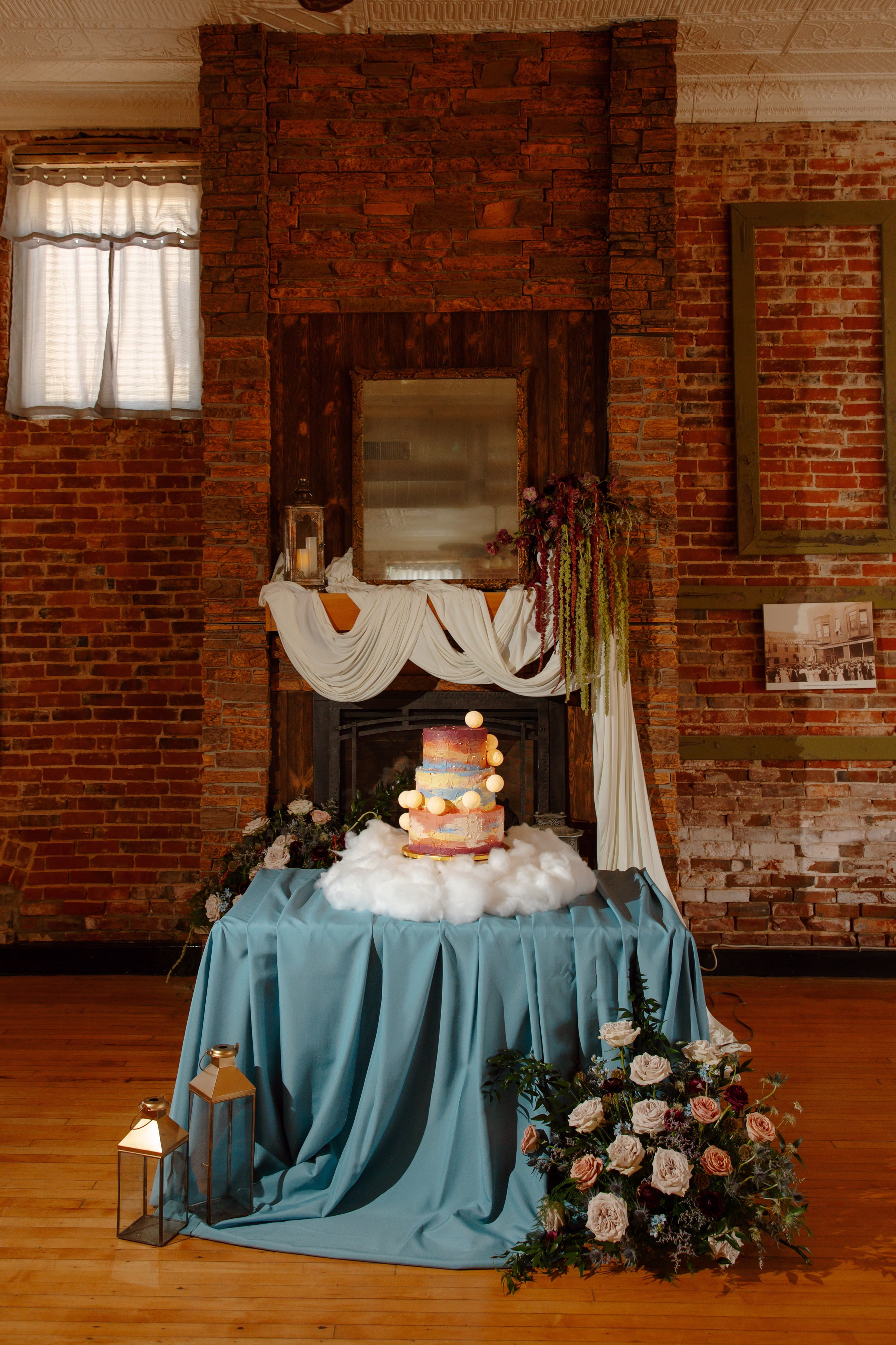 a celestial summer sunset wedding cake display by Cowboy David's Bakery with painted frosting and playful moons. The cake sits on faux clouds on a table with a blue tablecloth in front of the fireplace at B's Place Event Venue in West Salem, Wisconsin