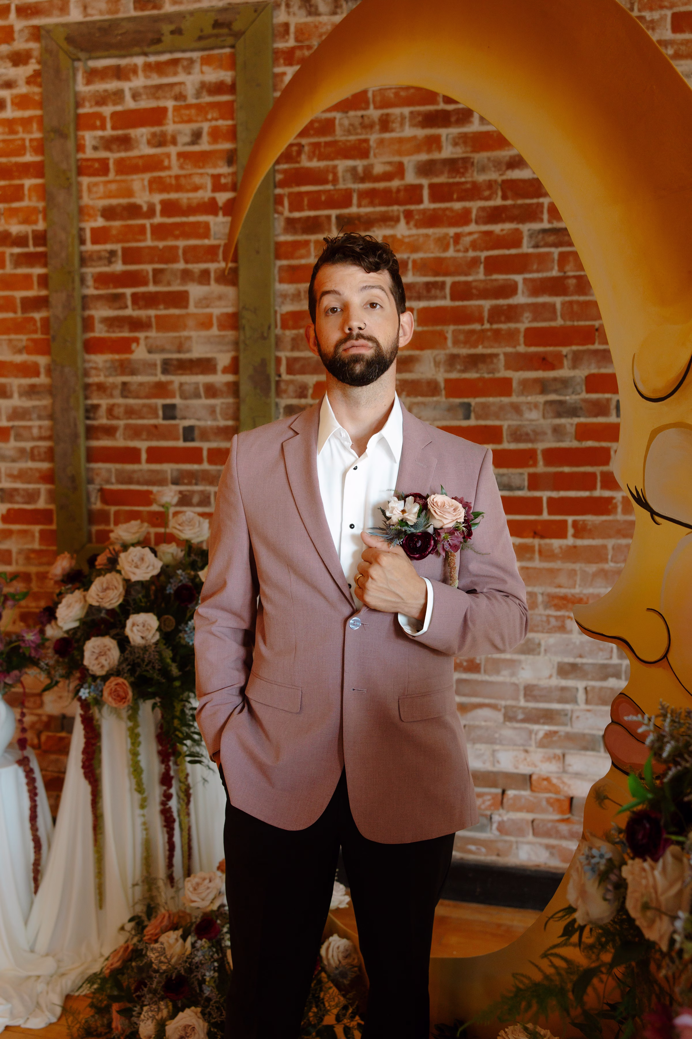 the groom stands at the ceremony backdrop with one arm holding the lapel of his dusty mauve suit jacket from Charlotte's Bridal & Formal Wear