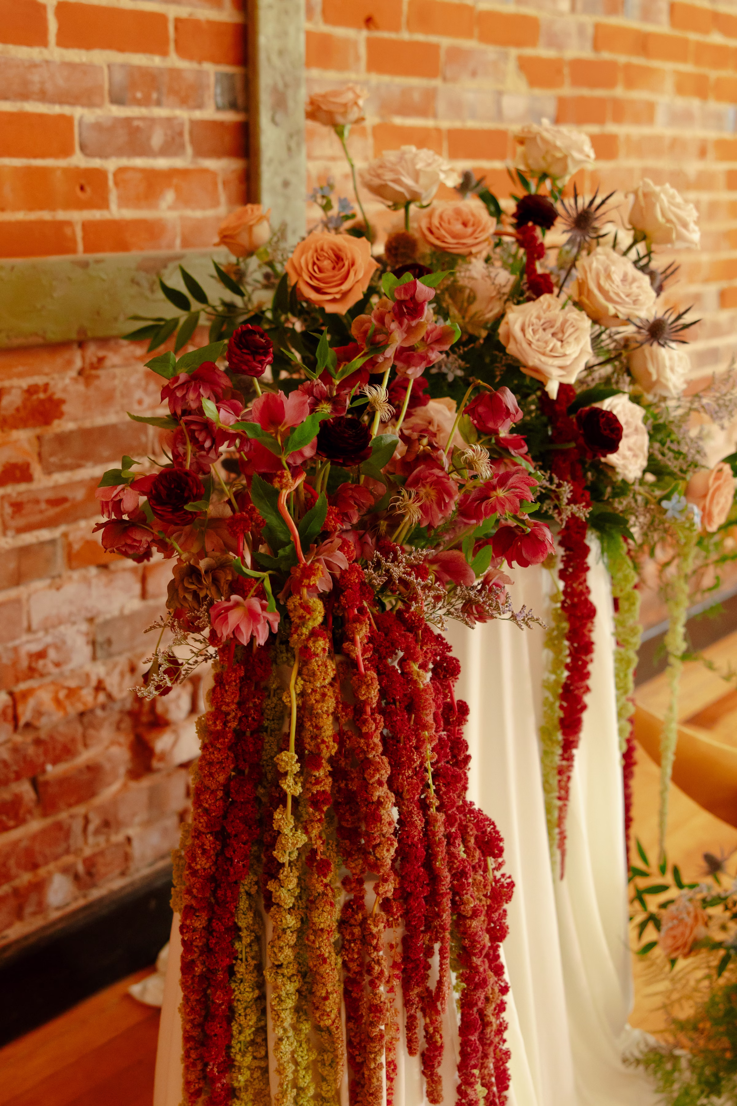 a close-up image of the ceremony florals by Bloom & Knot, heavily featuring white roses, blush roses, thistle, and red amaranths draping from the pedestal