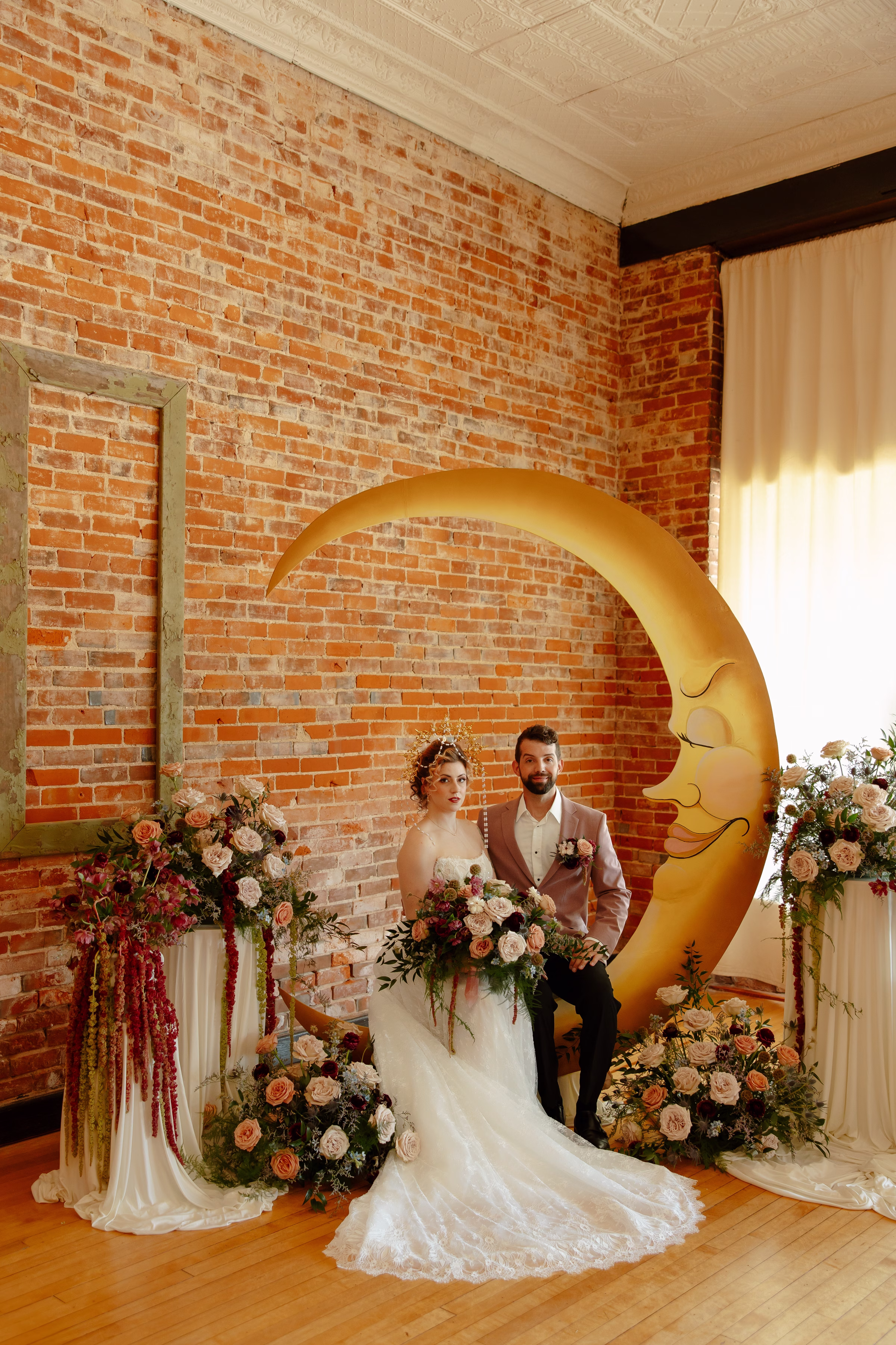 the bride and groom models sit in front of the smiling crescent moon ceremony backdrop, surrounded by florals