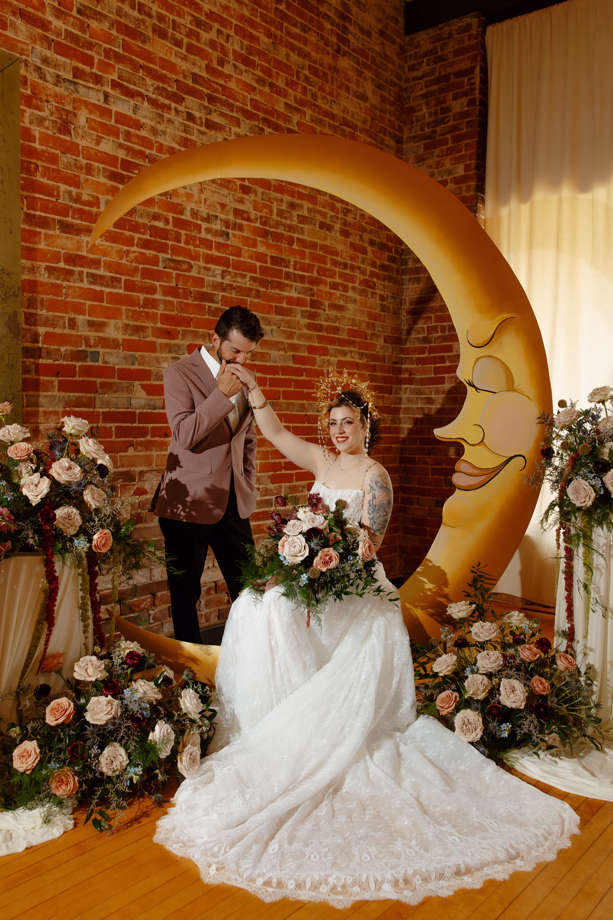 the groom kisses the bride's hand as she is seated at the crescent moon celestial wedding ceremony backdrop