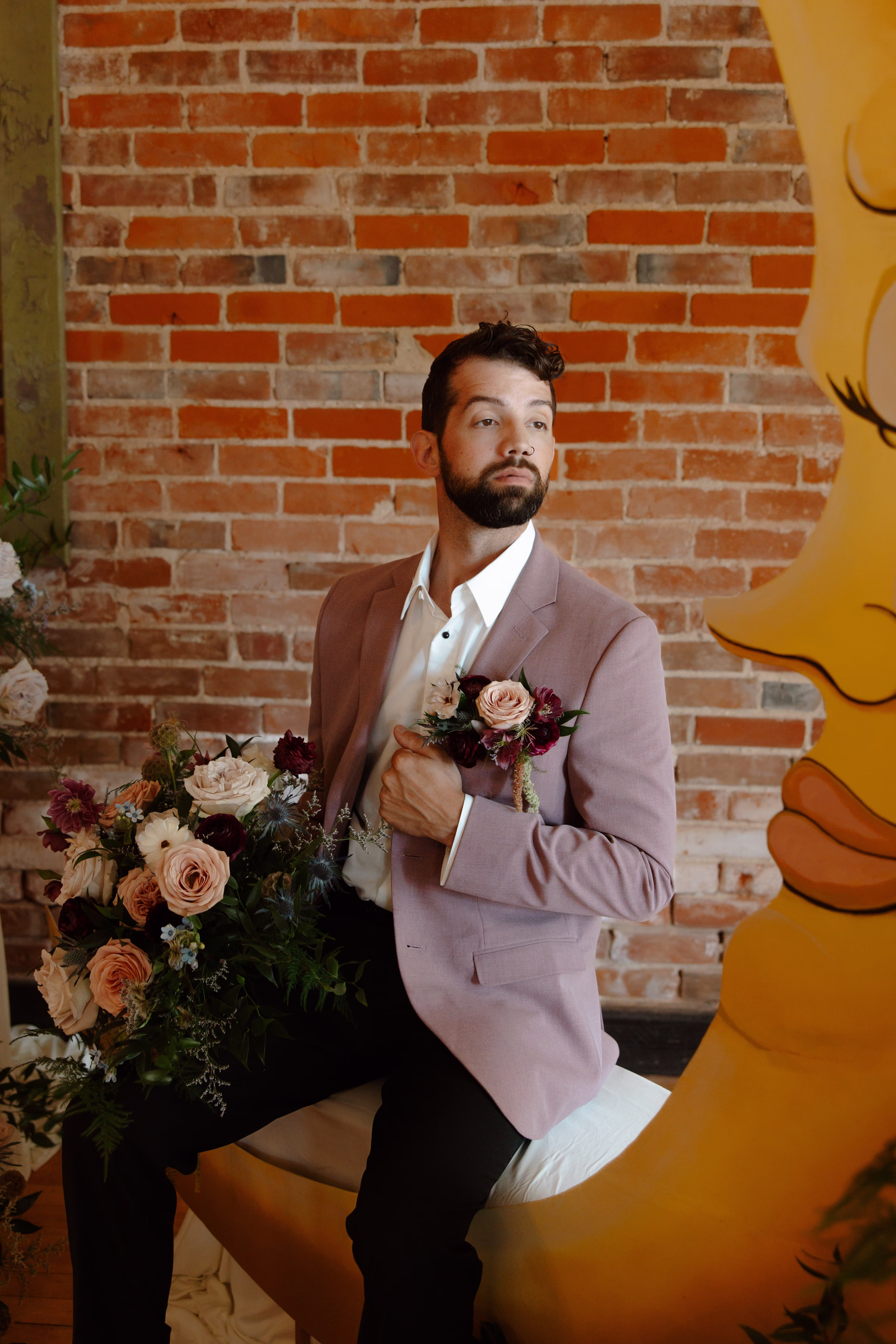 the groom sits at the ceremony setting with one arm holding the label of his dusty mauve suit jacket from Charlotte's Bridal & Formal Wear