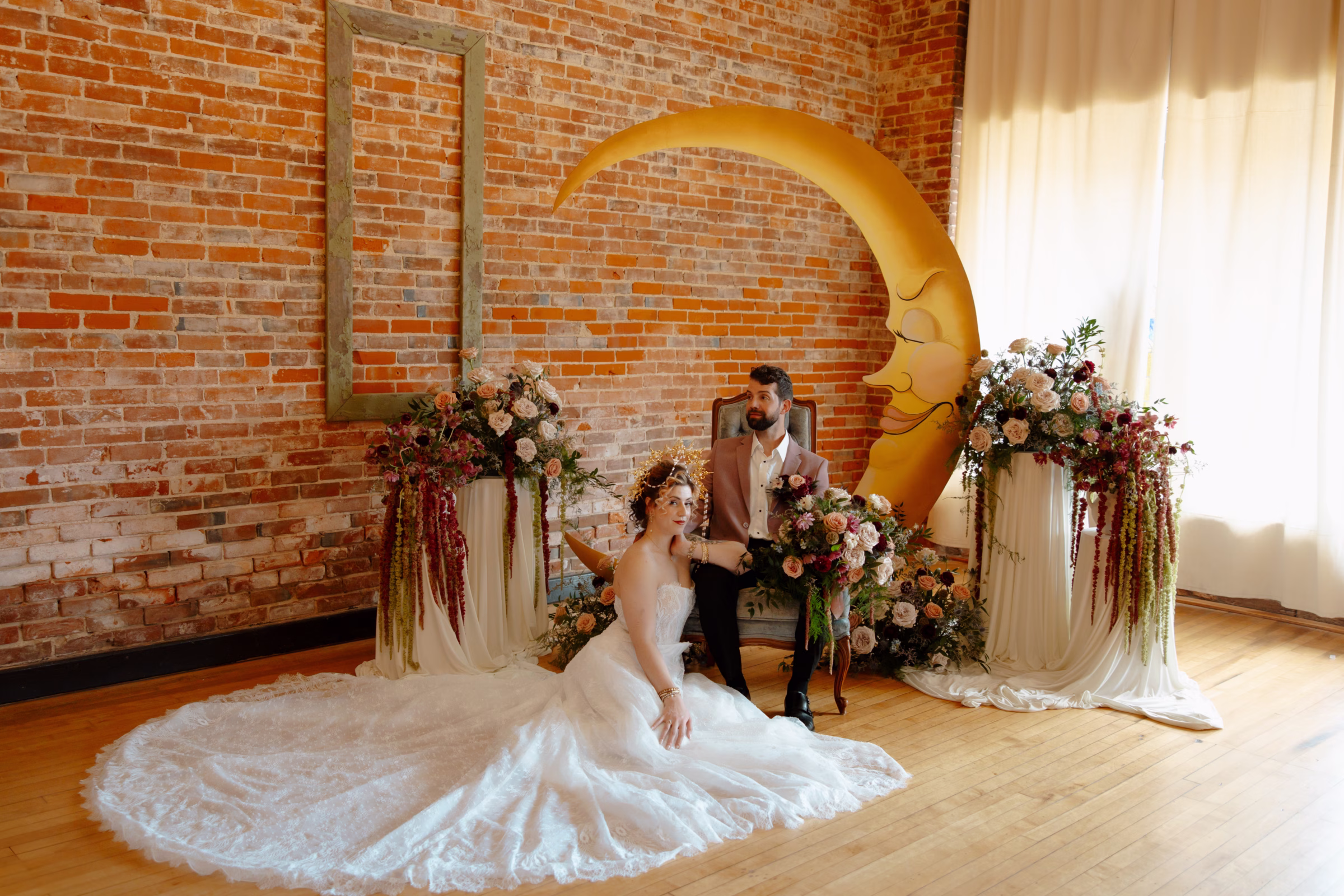 the bride and groom lounge at the ceremony backdrop. The groom is seated in a blue velvet chair, while the bride sits on the floor, showing off the train of her dress from Charlotte's Bridal & Formal Wear