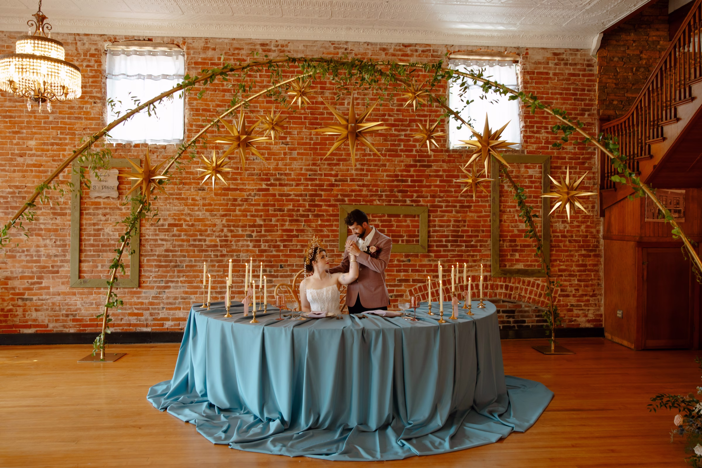 the couple poses beneath gold stars hung from an arch at the sweetheart table. The table is a half-circle covered with candles and a blue tablecloth cascading over the edge