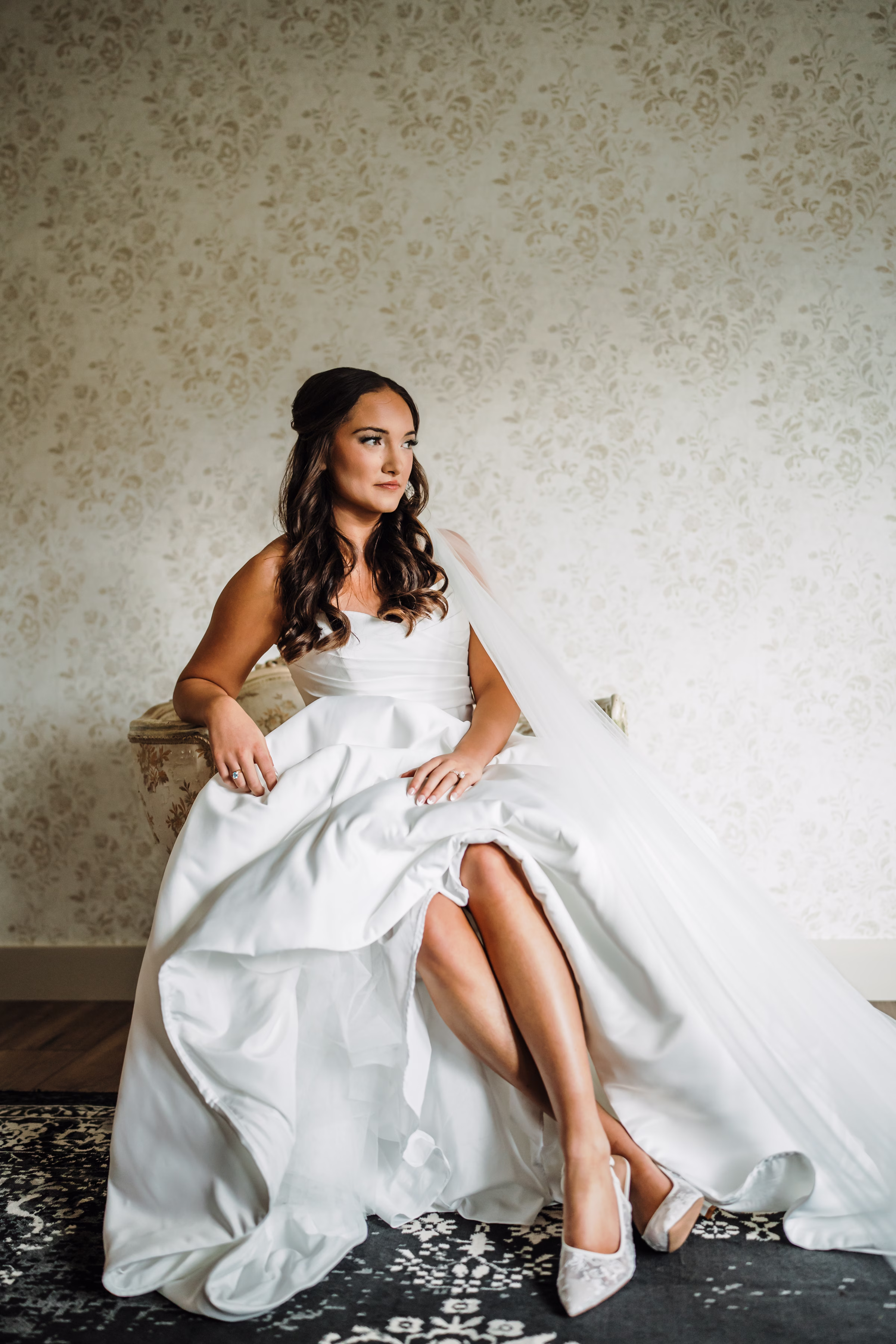scenic fall wedding winnebago springs minnesota bridal preparation 0224 - An Elegant Fall Wedding at Winnebago Springs | Emma & Tyler a bride is seated in her wedding dress against a wall with cream and taupe patterned wallpaper
