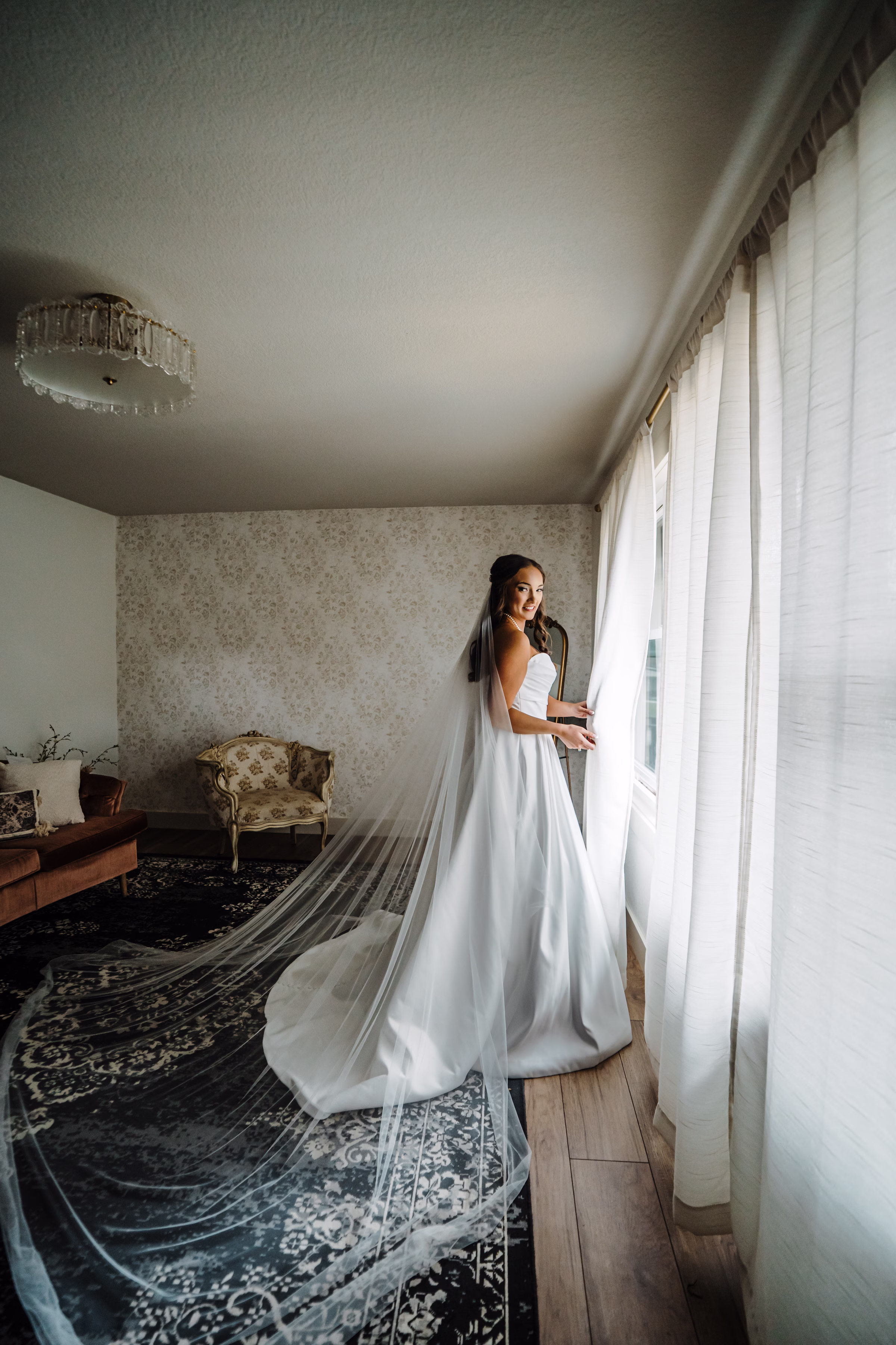 scenic fall wedding winnebago springs minnesota bridal preparation 3665 - An Elegant Fall Wedding at Winnebago Springs | Emma & Tyler a bride smiles by the window of her getting-ready room, showing off her delicate lace tulle cathedral-length veil