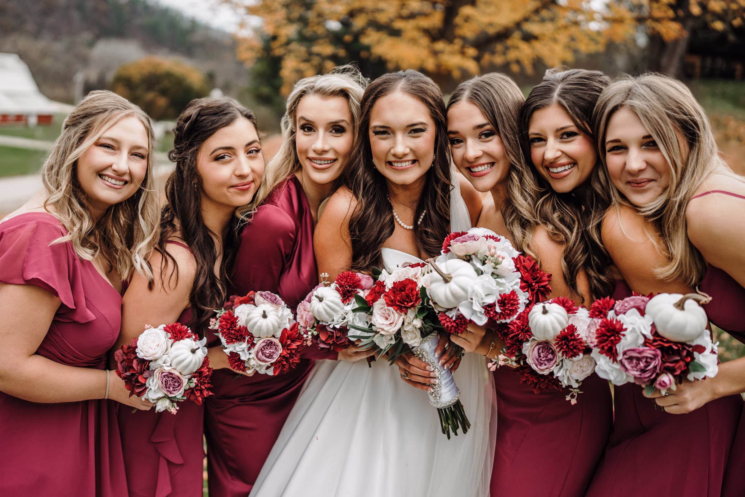 scenic fall wedding winnebago springs minnesota bridesmaids 1002 - An Elegant Fall Wedding at Winnebago Springs | Emma & Tyler a bride smiles, surrounded by her bridesmaids, all holding up bouquets. The florals are red, blush, white, and have small white pumpkins mixed in.