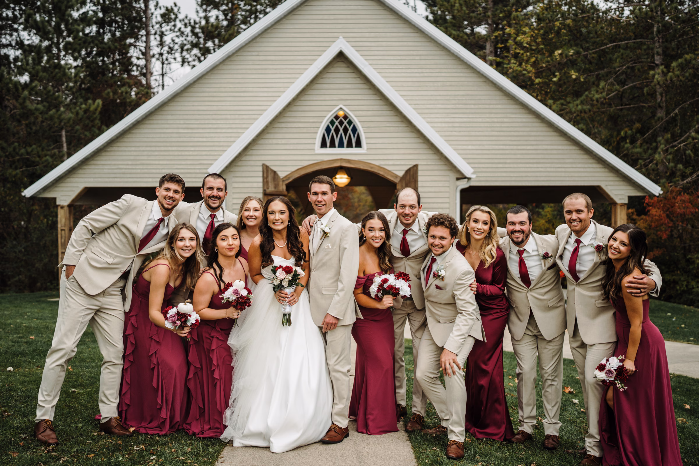 scenic fall wedding winnebago springs minnesota bridesmaids groomsmen 1161 - An Elegant Fall Wedding at Winnebago Springs | Emma & Tyler couple, Emma & Tyler, pose alongside their wedding party in an outdoor fall wedding photo. Bridesmaids are wearing burgundy dresses and groomsmen are wearing taupe suits with brown shoes, white dress shirts, and burgundy ties and pocket squares