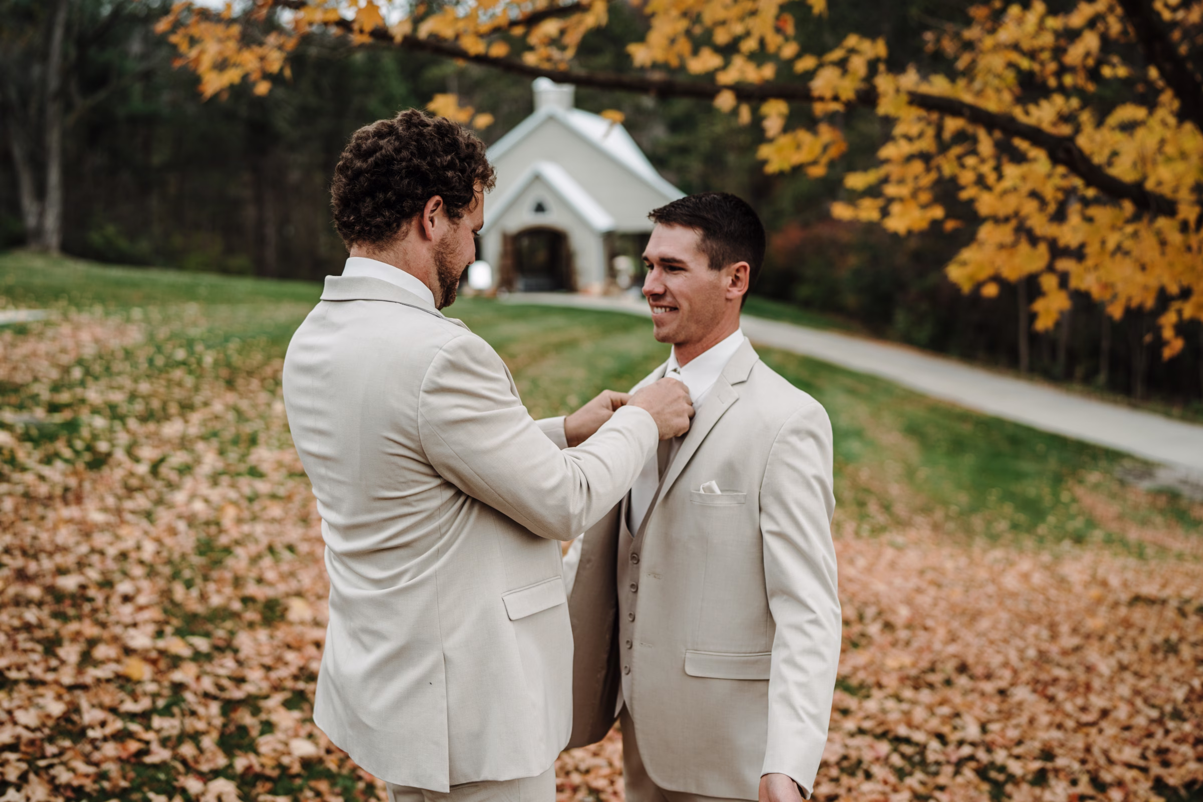 scenic fall wedding winnebago springs minnesota groom preparation 3760 - An Elegant Fall Wedding at Winnebago Springs | Emma & Tyler a groomsman helps secure a groom's tie, as they stand outside Winnebago Springs' air chapel surrounded by fall colors