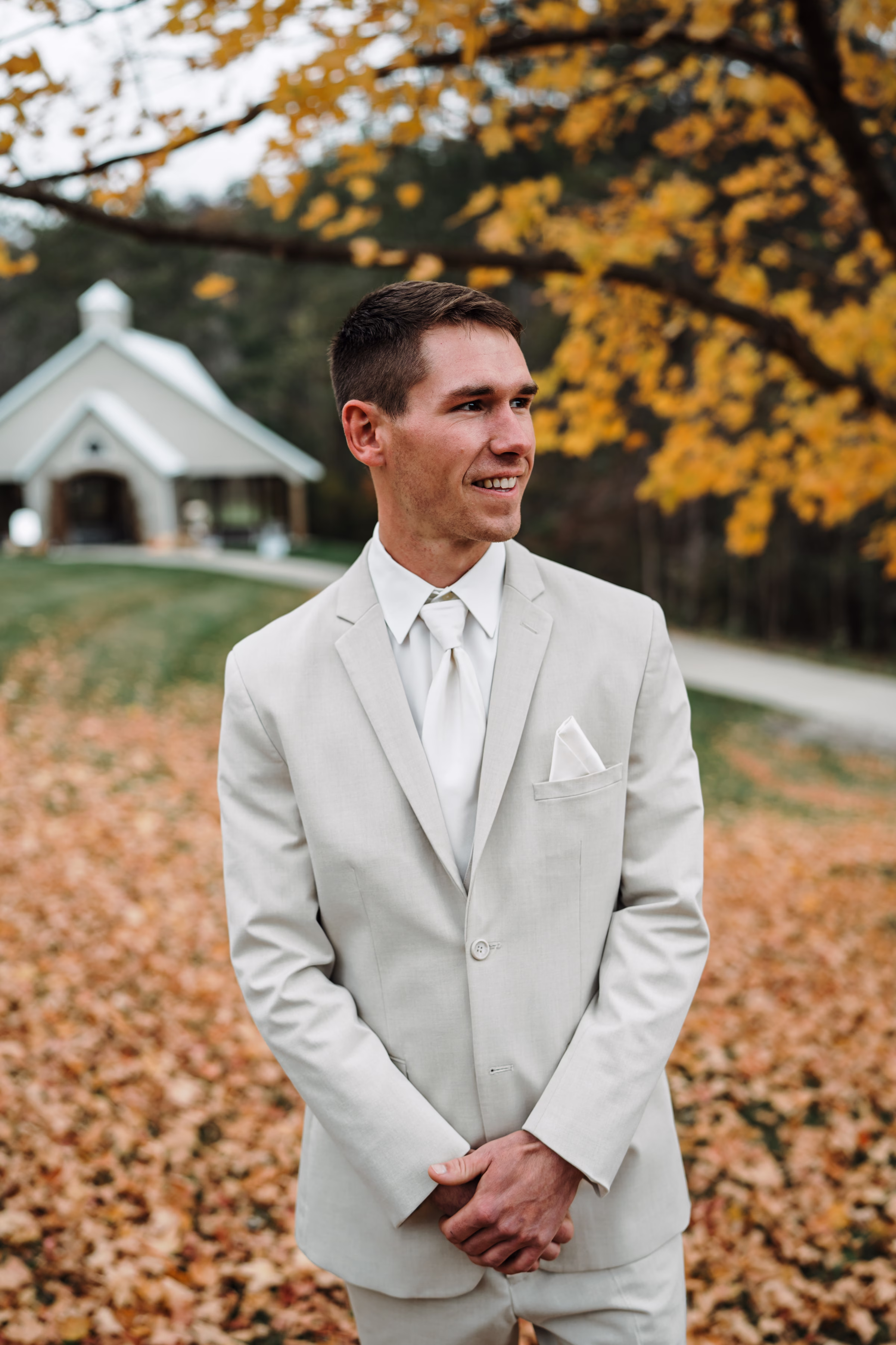 scenic fall wedding winnebago springs minnesota groom preparation 3791 - An Elegant Fall Wedding at Winnebago Springs | Emma & Tyler a groom smiles and looks off into the distance, surrounded by fall foliage. He is wearing a light taupe or gray suit with white dress shirt, white tie, and white pocket square
