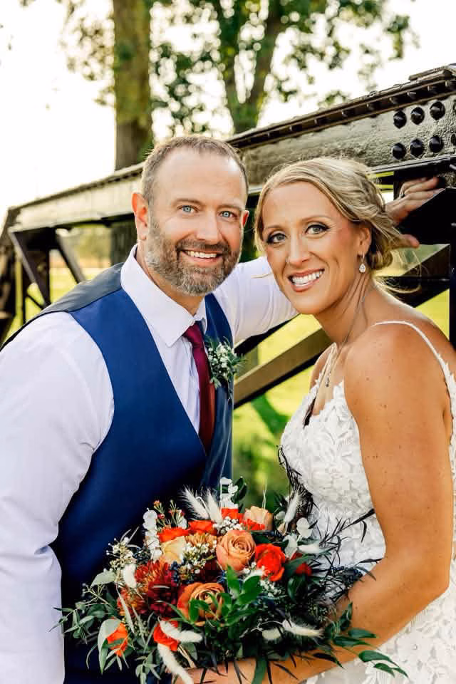 A smiling bride and groom lean in together outdoors by a black metal structure, highlighting her flawless, natural-looking wedding spray tan by Bronzd by Bre.