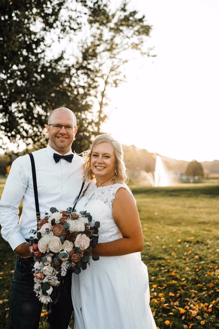 A smiling bride and groom pose at golden hour on a grassy lawn with a fountain in the background, showcasing her sun-kissed, natural-looking spray tan by Bronzd by Bre.