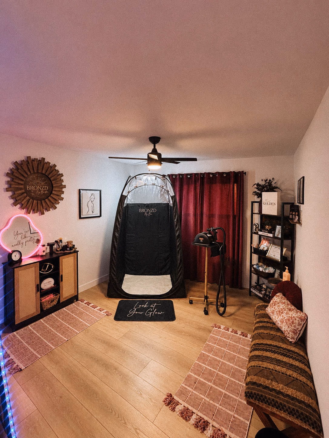 The complete upscale interior of Bronzd by Bre, featuring a private airbrush tent, professional tanning machine, cozy waiting bench, and a pink neon sign.