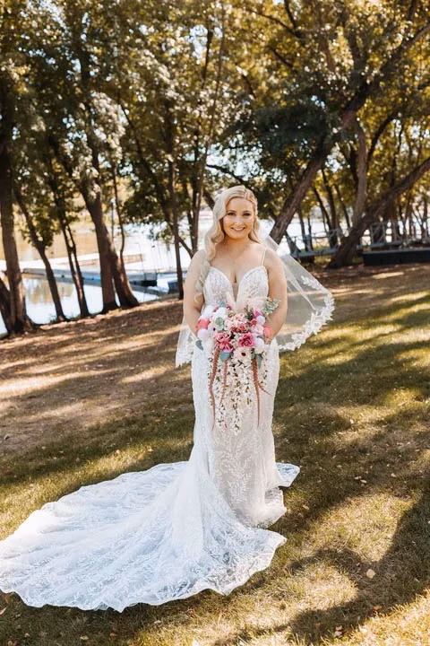 A smiling bride poses outdoors in a lace wedding gown with a flowing train and cascading pink bouquet, showcasing an effortlessly blended, natural-looking spray tan by Bronzd by Bre.