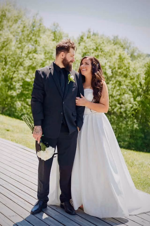An outdoor wedding portrait of a couple looking at each other, showcasing a groom in a sleek all-black suit and a bride with a beautiful sun-kissed glow by Bronzd by Bre in La Crosse County.